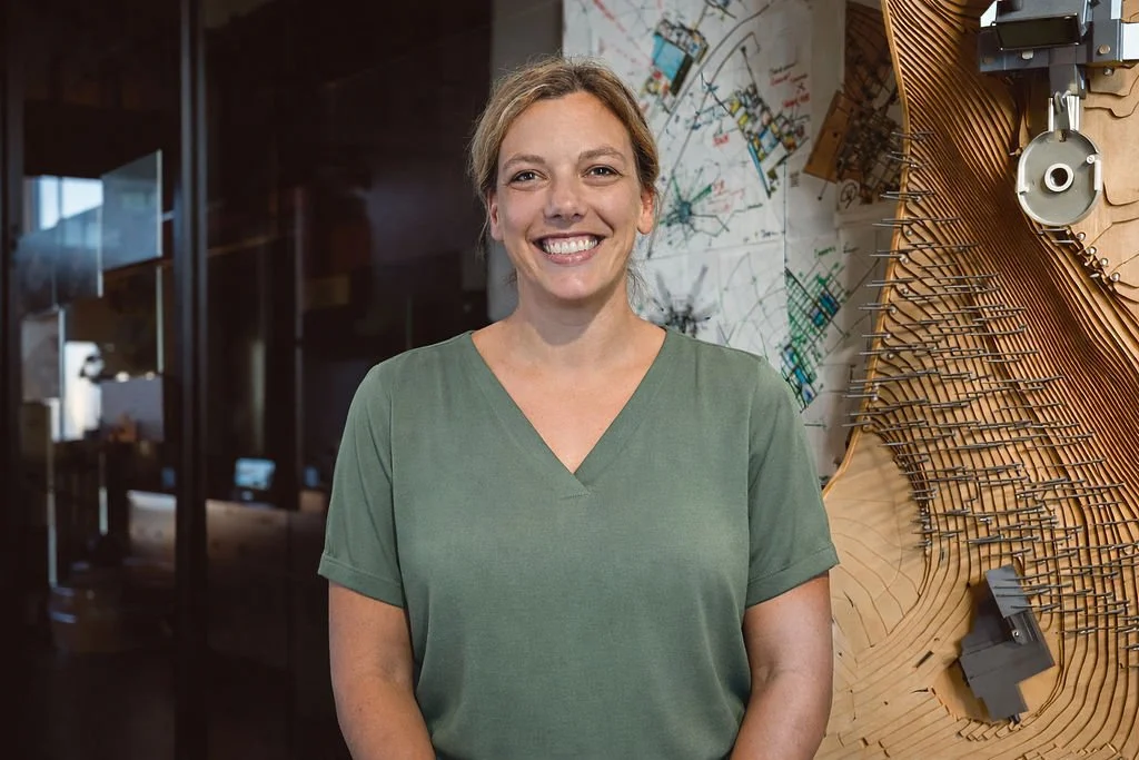 A woman wearing a green scrub top standing in front of a wall with mechanical and architectural diagrams and wooden structures.