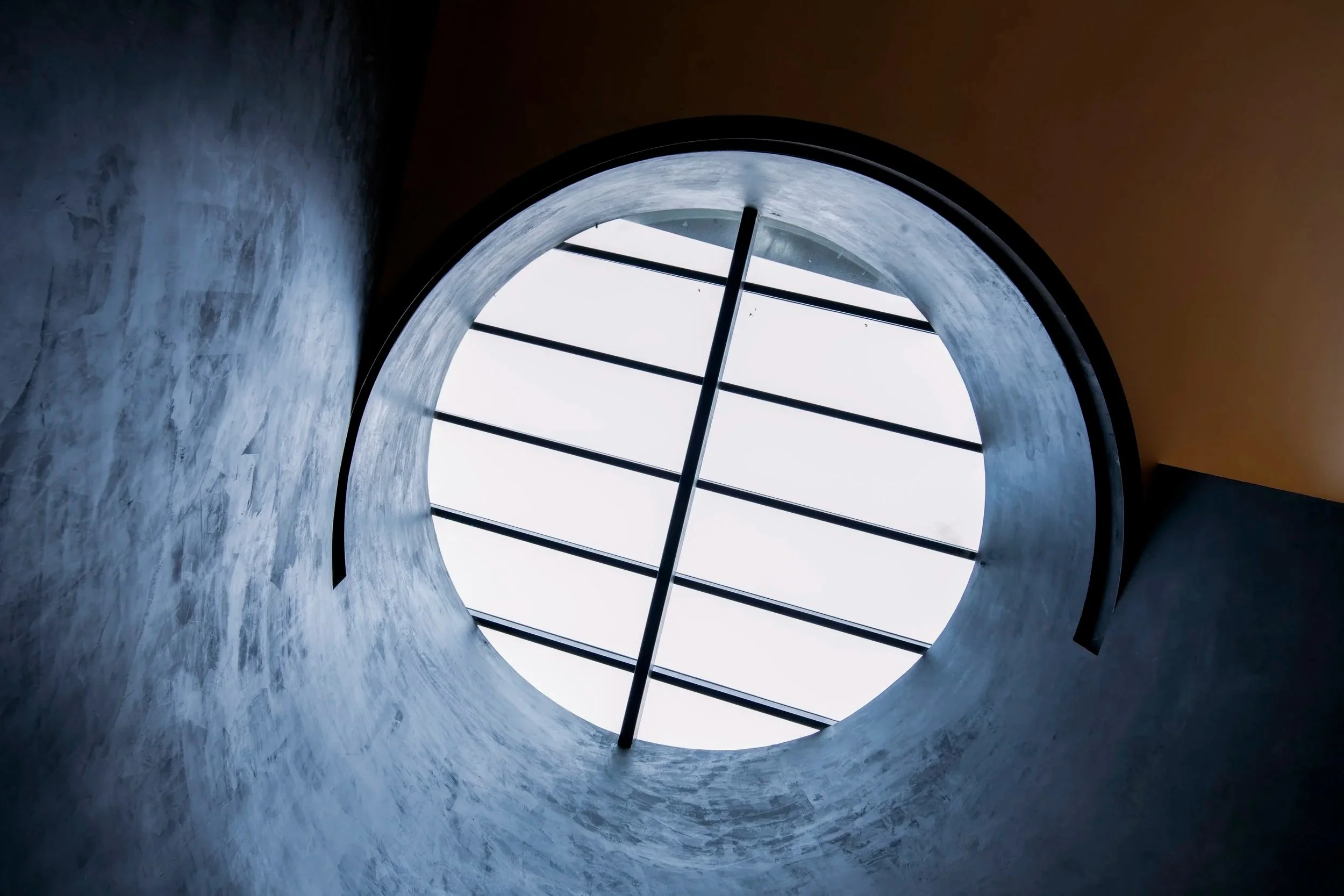 View of a circular skylight from below, showing the sky and grid-like metal bars through the opening.