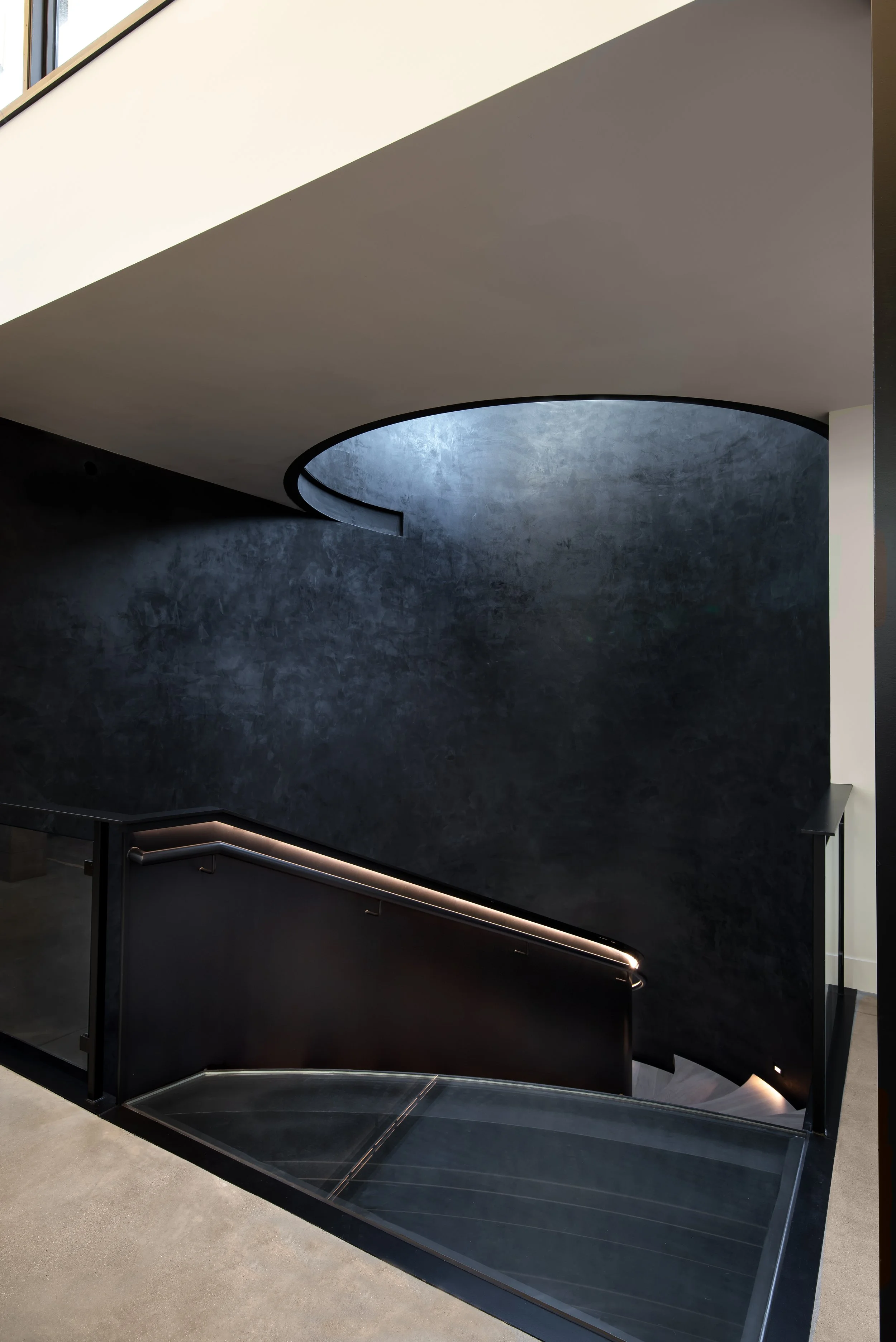 Modern interior staircase with black walls and railing, illuminated by subtle lighting, leading down from a white ceiling with geometric cutout holes.