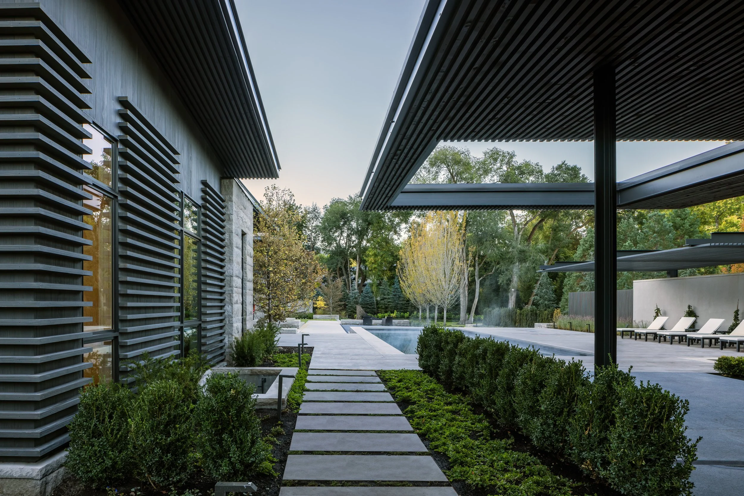 Modern backyard with a pathway, pool, lounge chairs, and trees, surrounded by contemporary architecture with metal and stone accents.