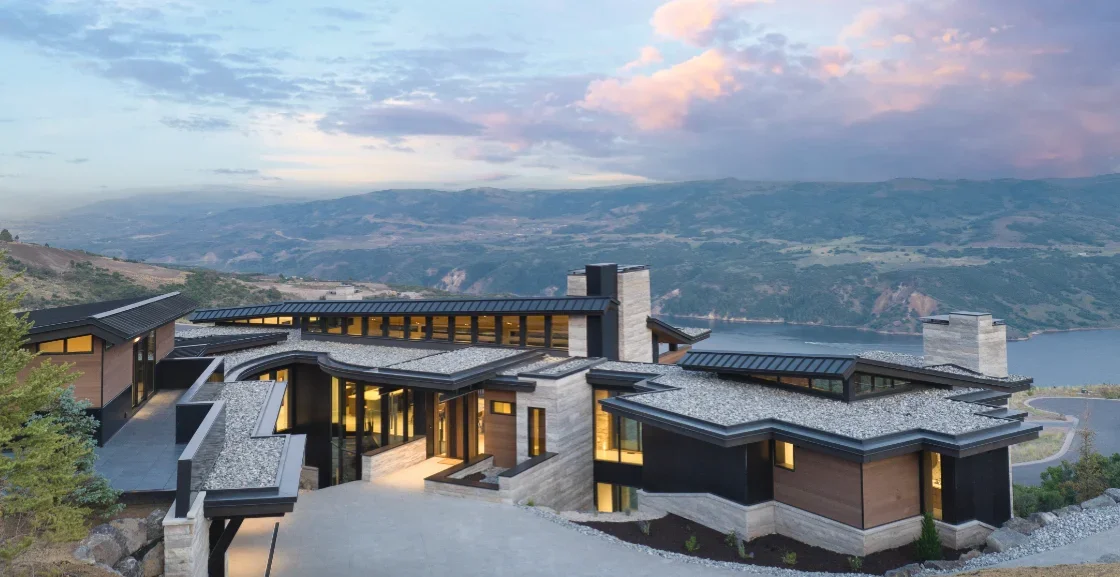 Modern house on hillside overlooking a body of water with mountains in the background, during sunset.