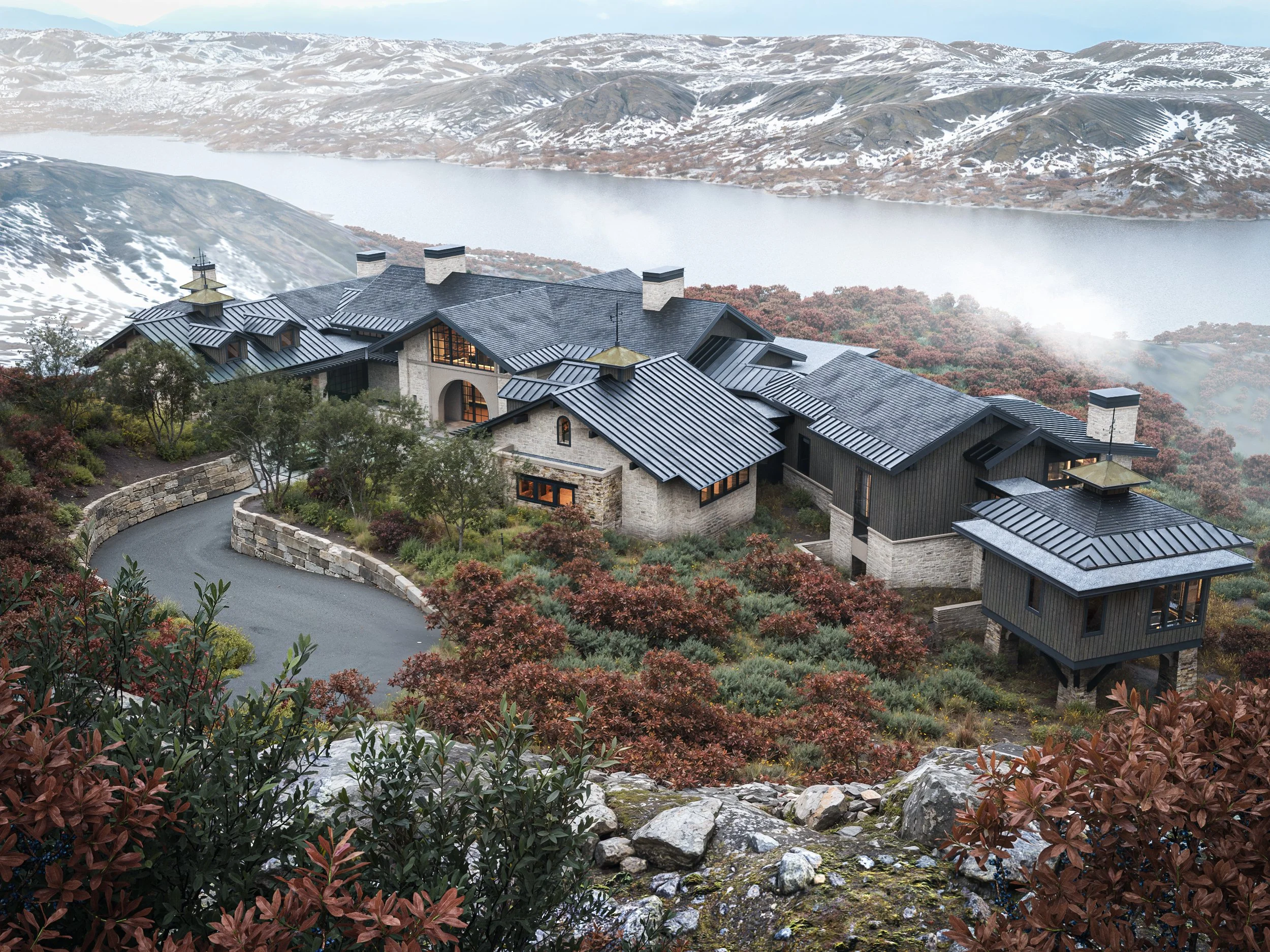 A large house with multiple sections, stone and dark wood exterior, surrounded by trees and shrubs, located on a hillside overlooking a lake and mountains in the background.