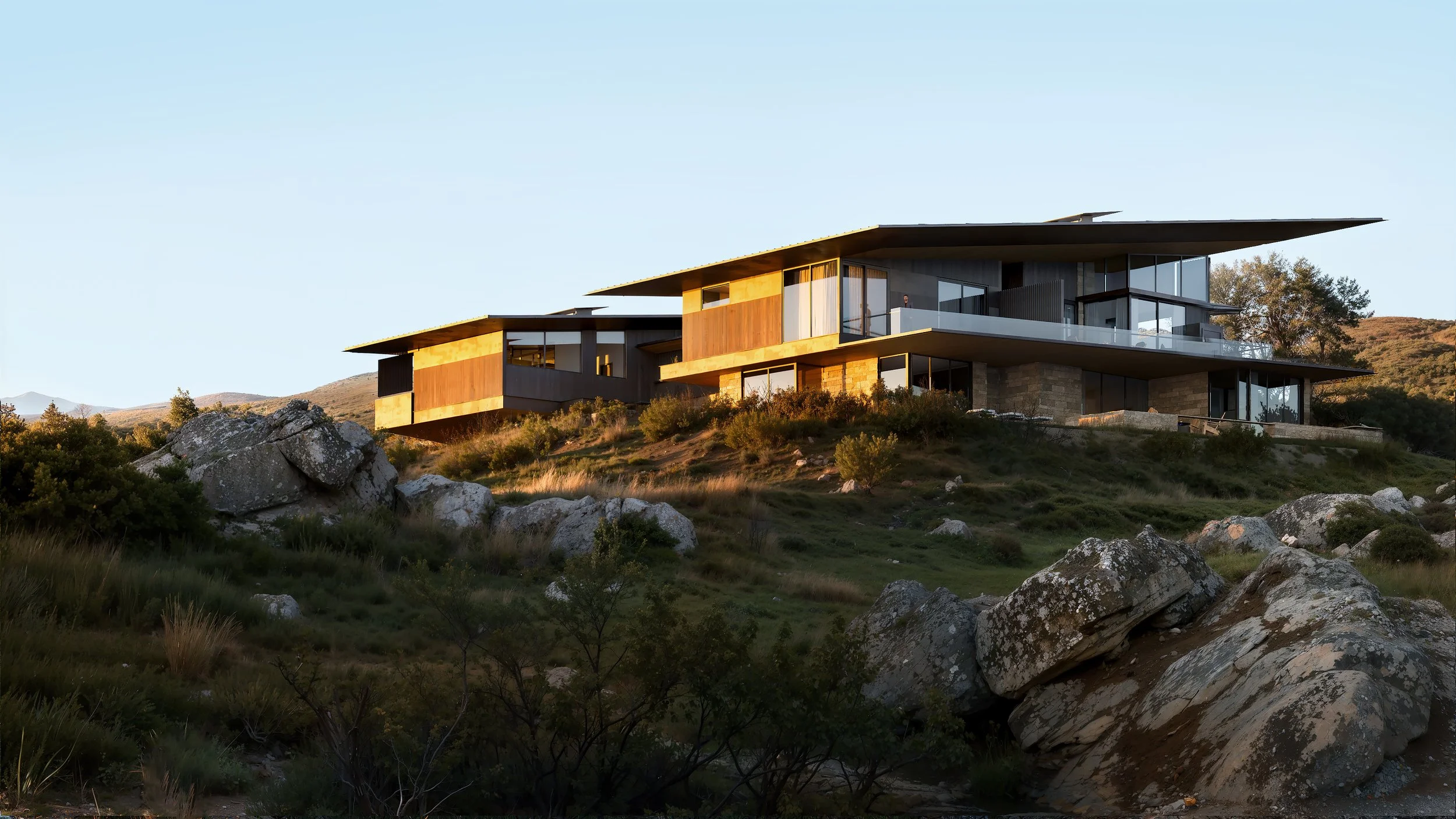 Modern house with large glass windows and flat roofs, situated on a hillside with rocks and vegetation, during sunset or sunrise.