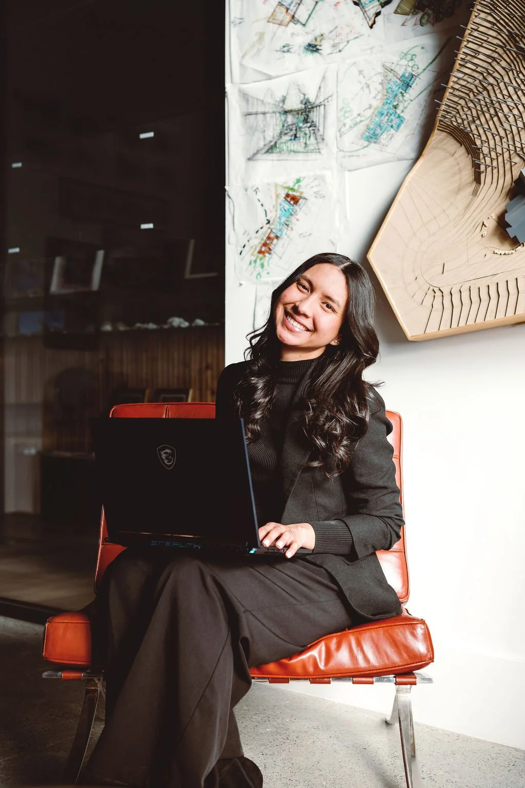 A smiling woman in a black suit sitting on an orange chair with a laptop on her lap in an office or workspace.