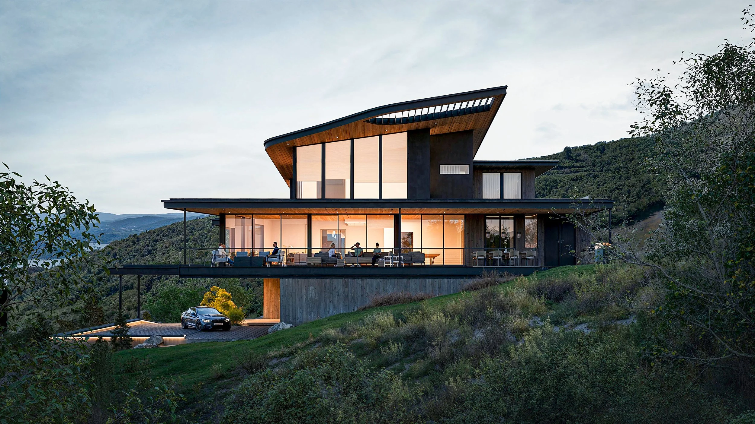 Modern multi-story house on a hillside with large glass windows, a balcony with people sitting, and a driveway with a car, surrounded by greenery and mountains in the background during dusk.