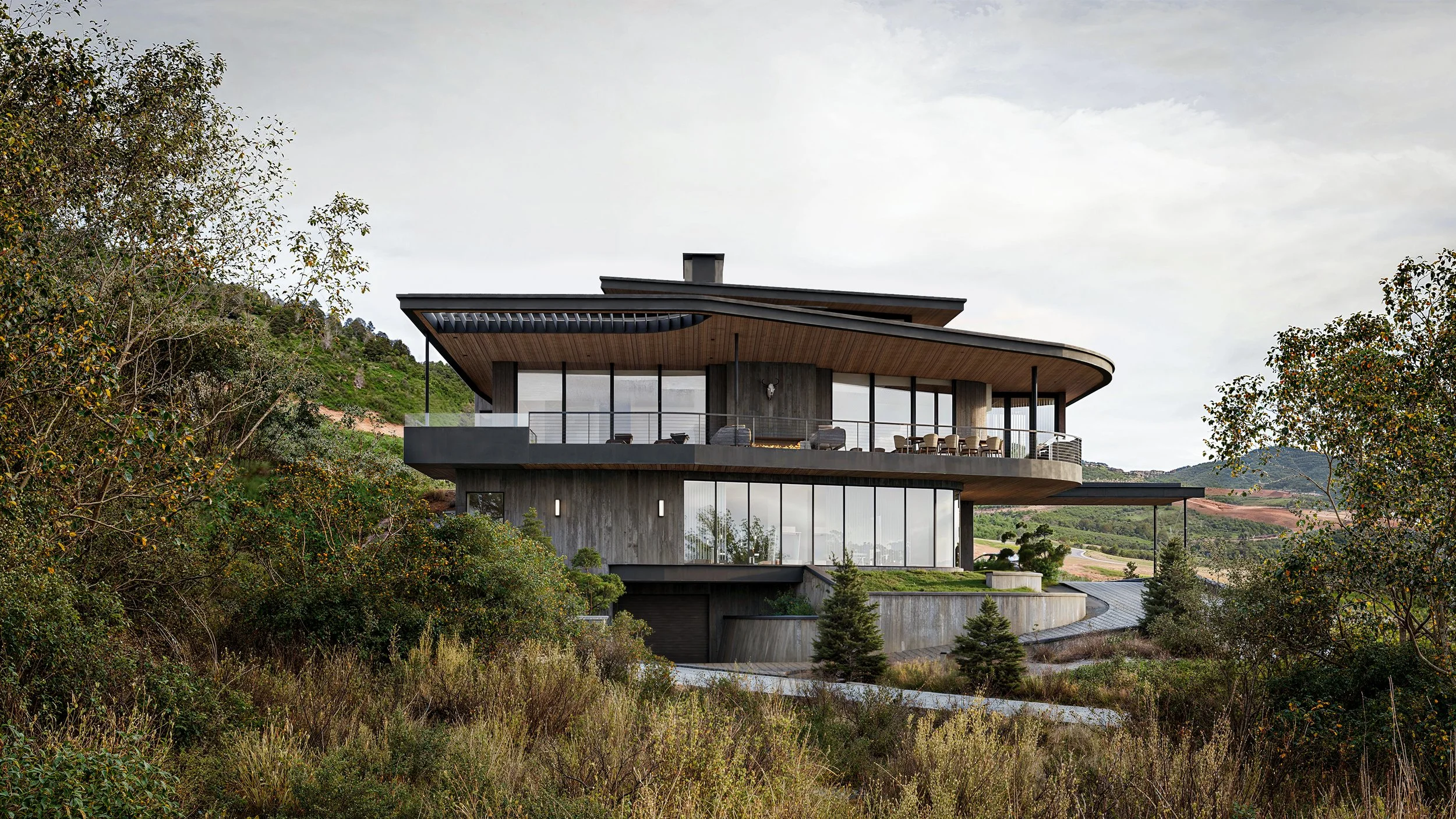 Modern house with large glass windows and an expansive balcony, situated on a hillside surrounded by trees and greenery.