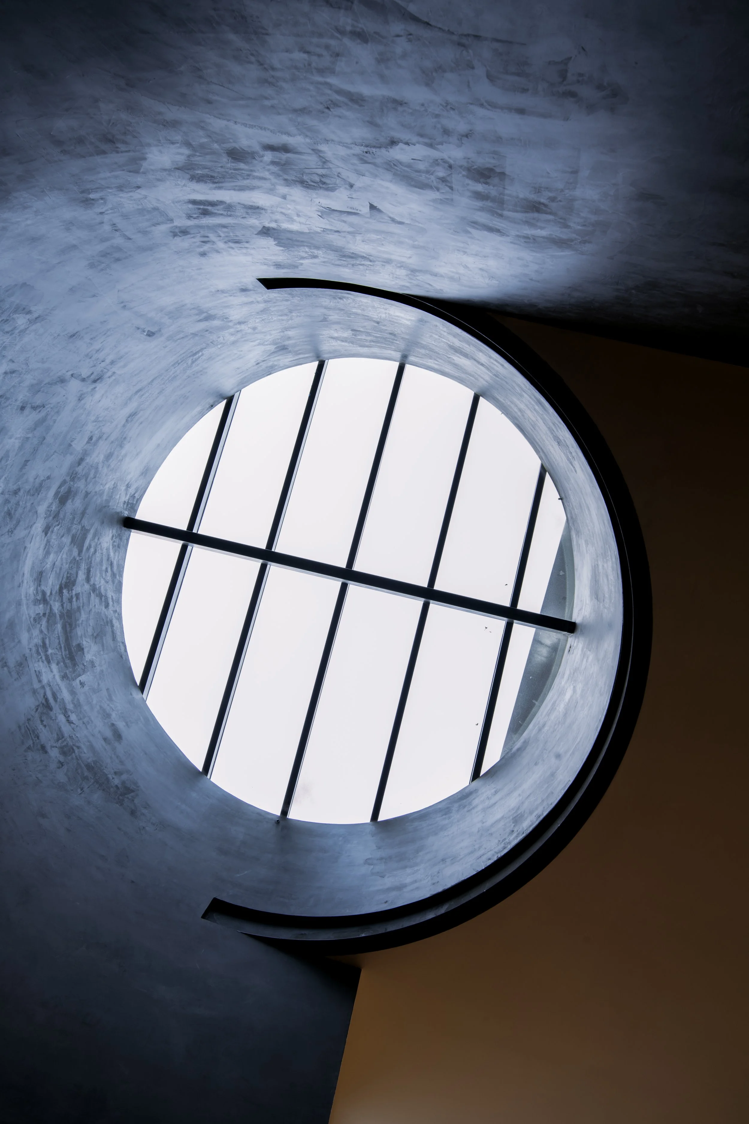 Looking up through a circular skylight with black metal bars, showing a cloudy sky.
