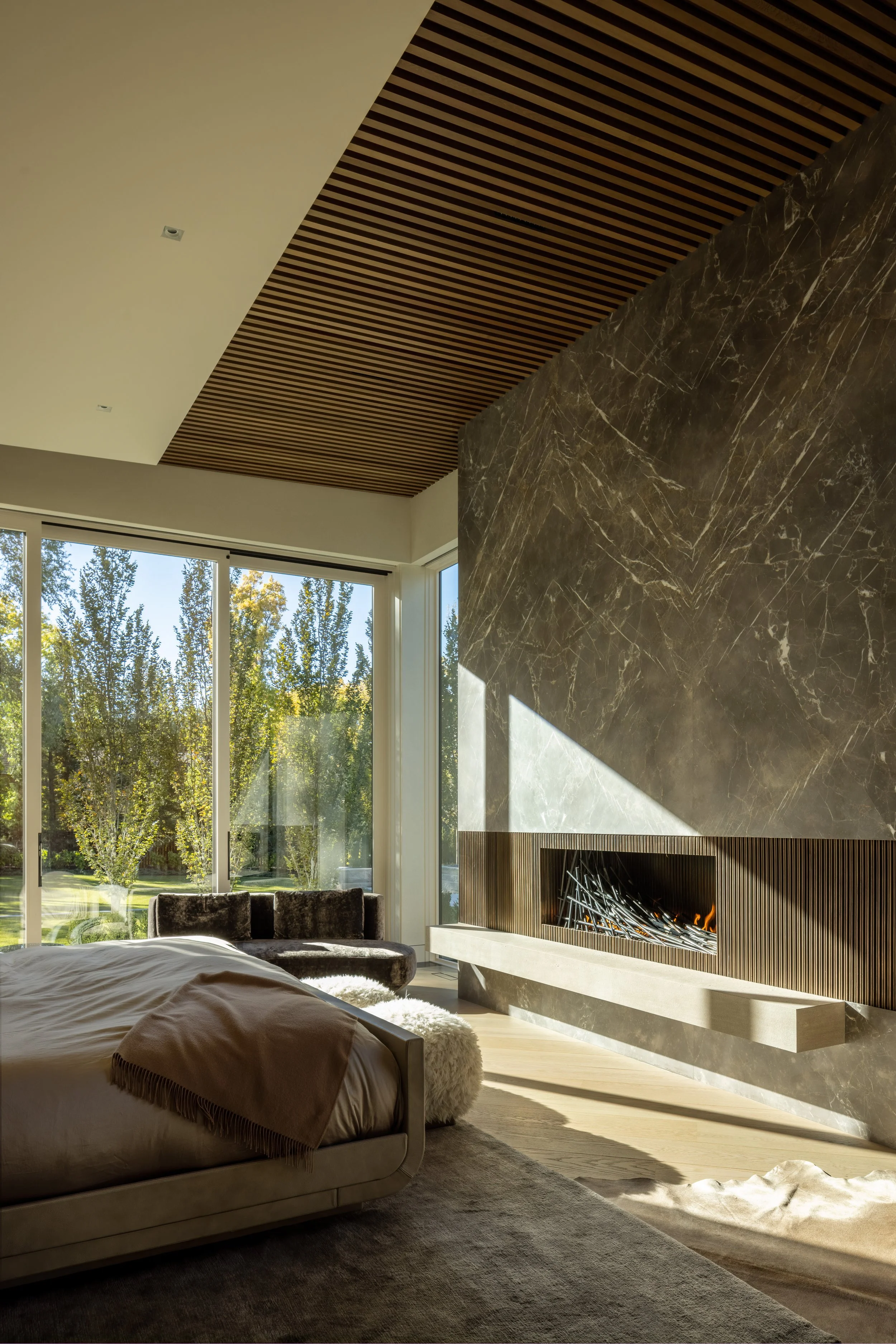 Modern living room with large floor-to-ceiling windows, a wall fireplace, a bed, and a view of trees outside.