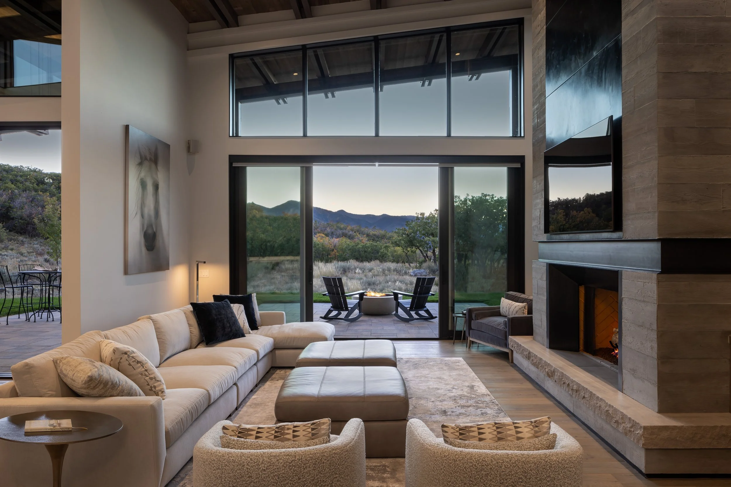 Modern living room with large glass windows showing mountainous landscape and outdoor seating area with fire pit