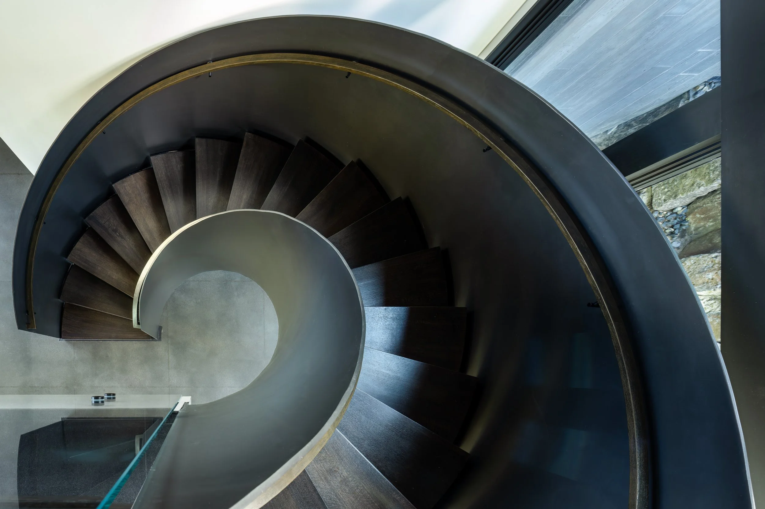 Top-down view of a winding spiral staircase with dark wooden steps and a black railing, next to a glass wall showing outdoor scenery.