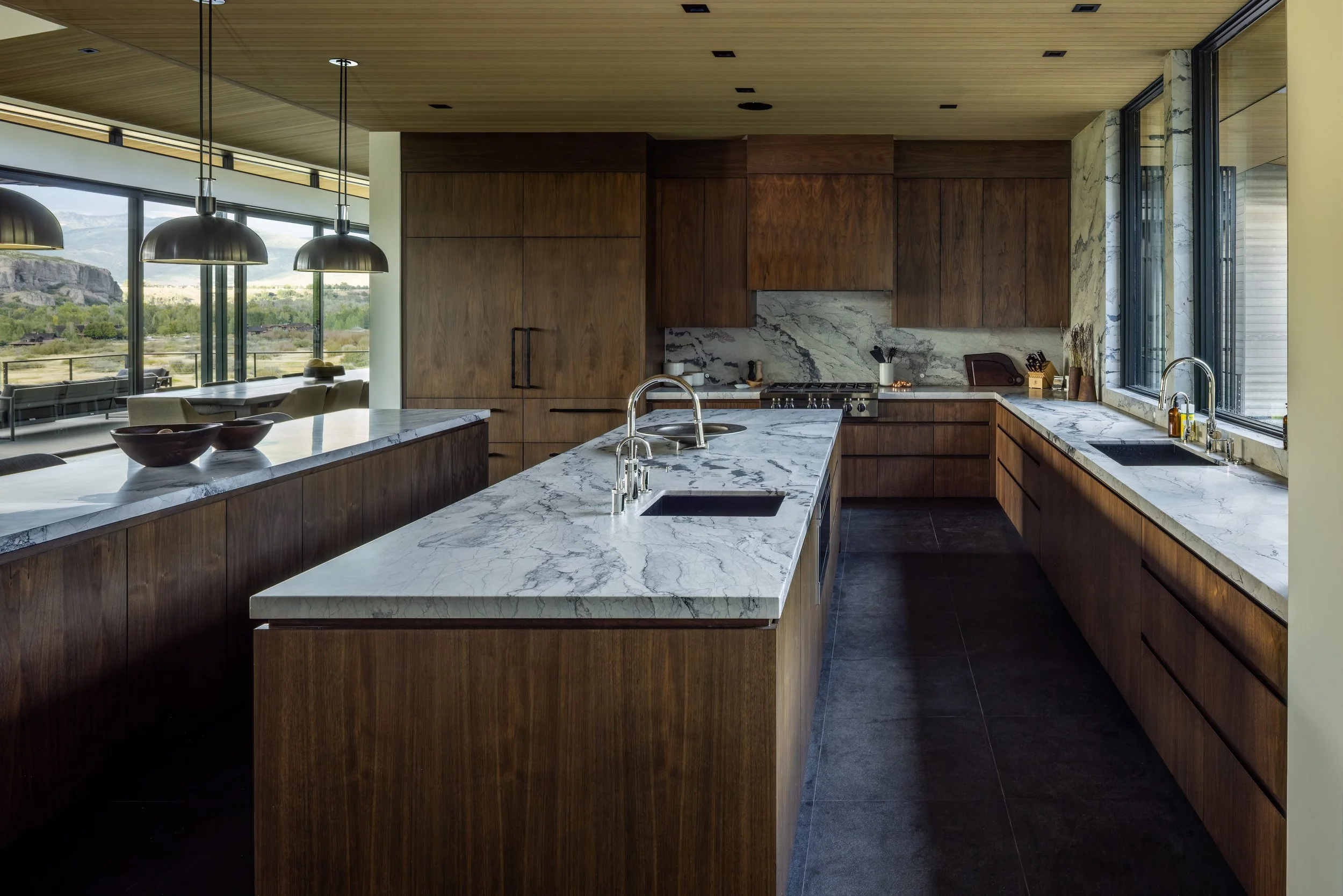Modern kitchen with wooden cabinets and marble countertops, large windows overlooking a landscape, and black pendant lights.
