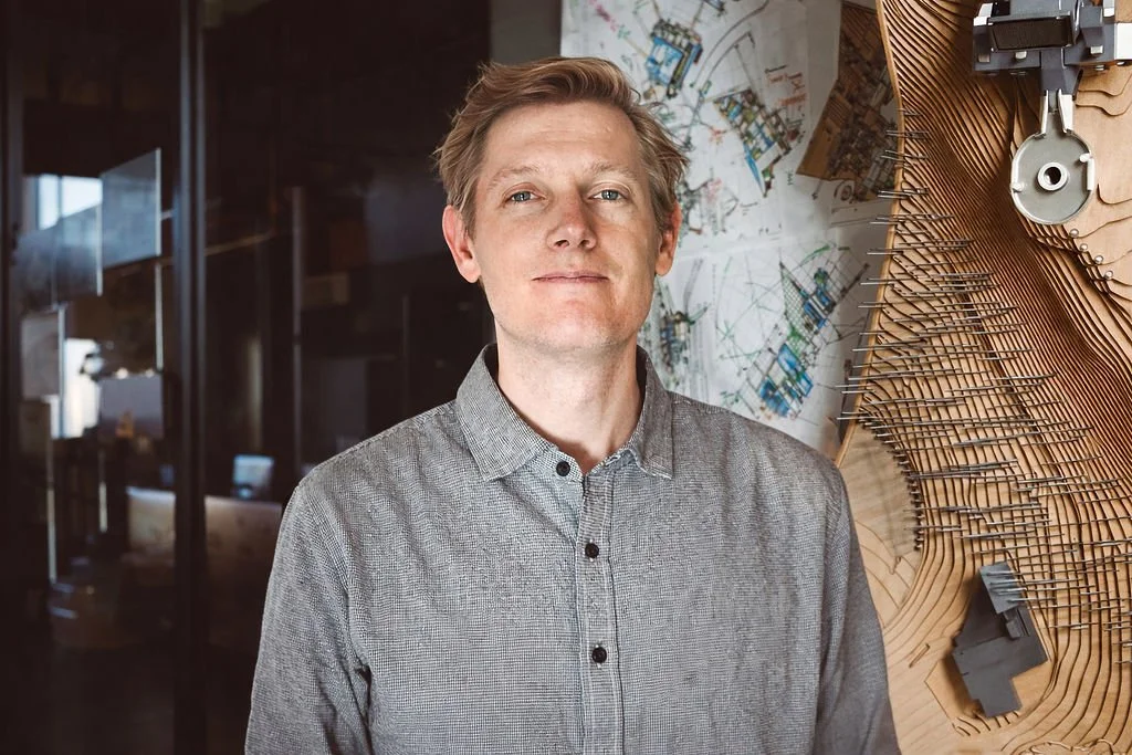 A man with blond hair wearing a gray button-up shirt standing in front of architectural or engineering plans and a large wooden model or sculpture in an office environment.