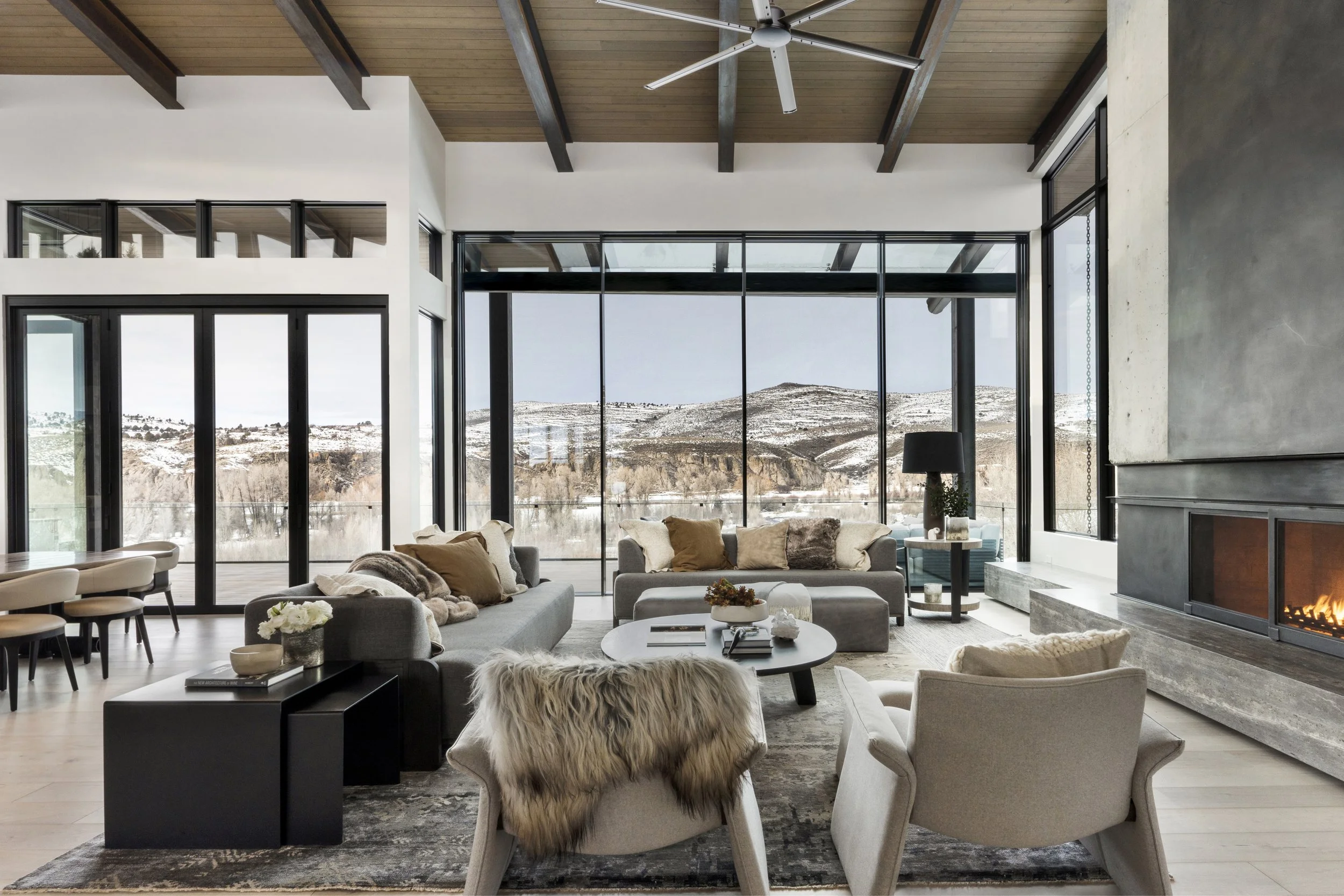Modern living room with large glass windows offering a scenic mountain view, featuring a fireplace, beige armchairs, a gray sofa with pillows, and a round coffee table, in a high-ceiling space with wood and concrete accents.