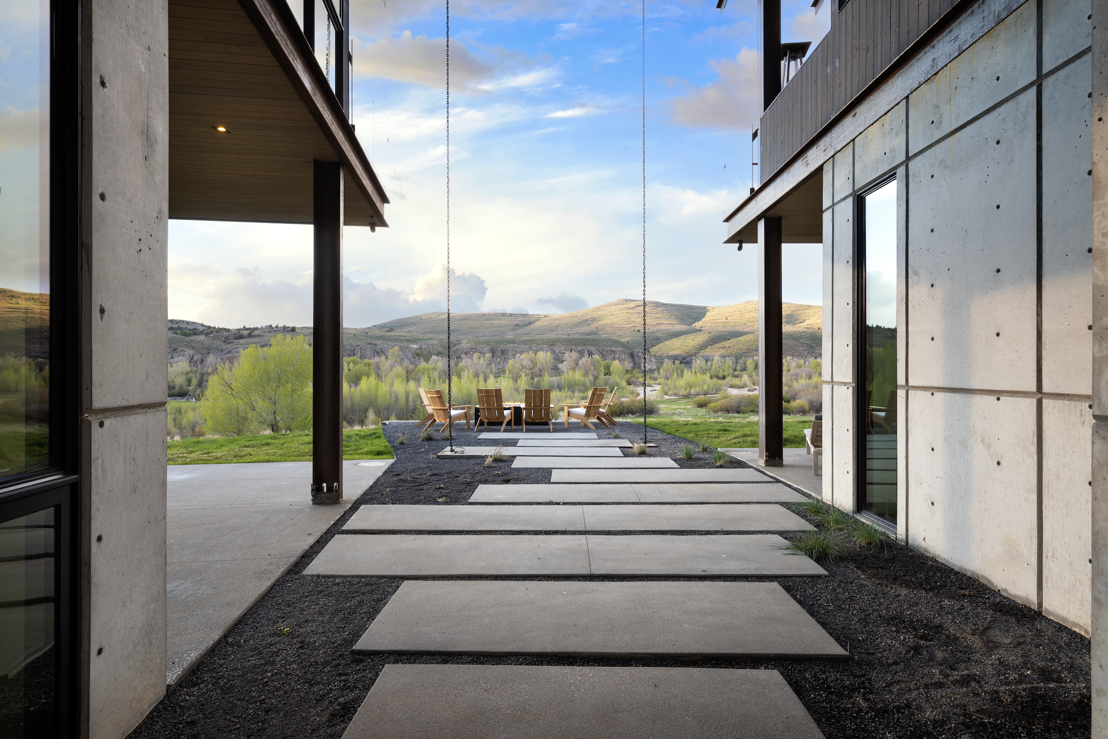 Modern house patio with concrete stepping stones leading to a view of green landscape and hills, outdoor wooden chairs arranged around a table.