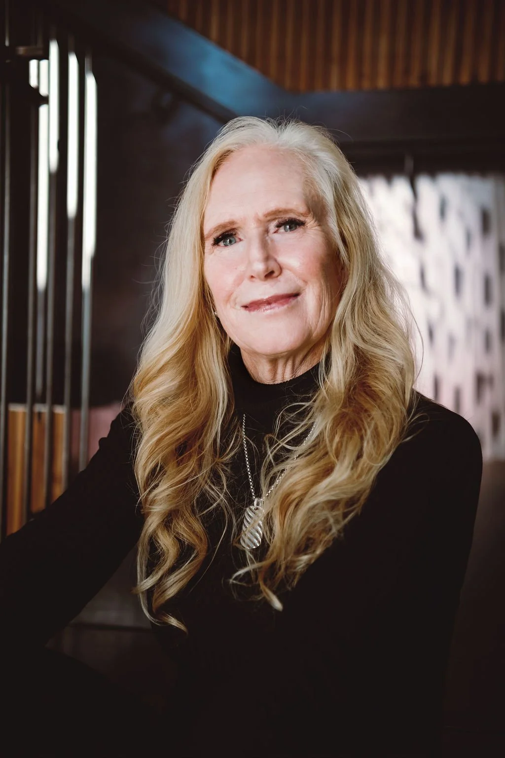 A portrait of an elderly woman with long, wavy blonde hair, wearing a black top and a silver necklace, smiling gently indoors.
