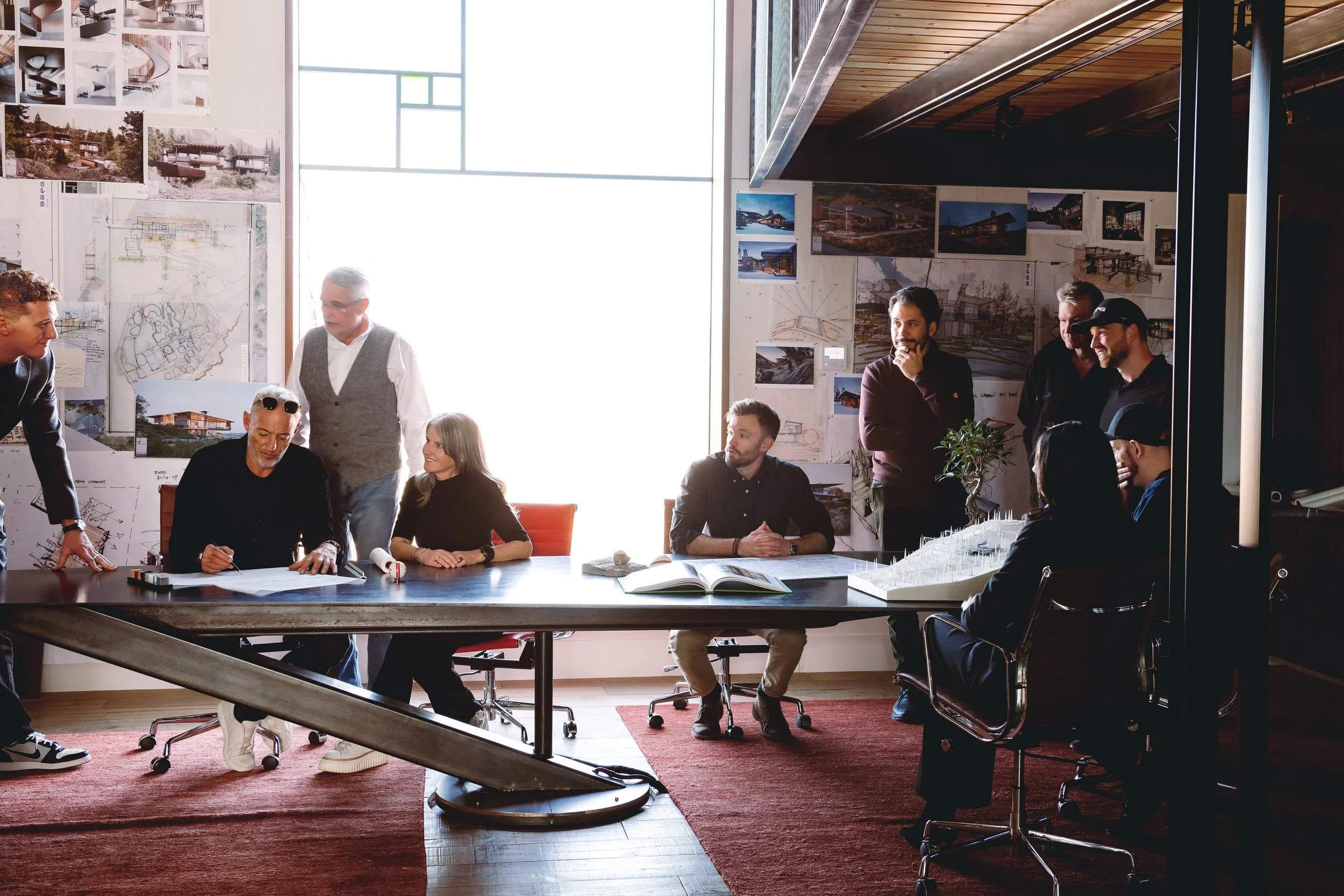 Group of people gathered around a large table in a modern office or conference room, discussing and collaborating with architectural plans and photos on the wall.