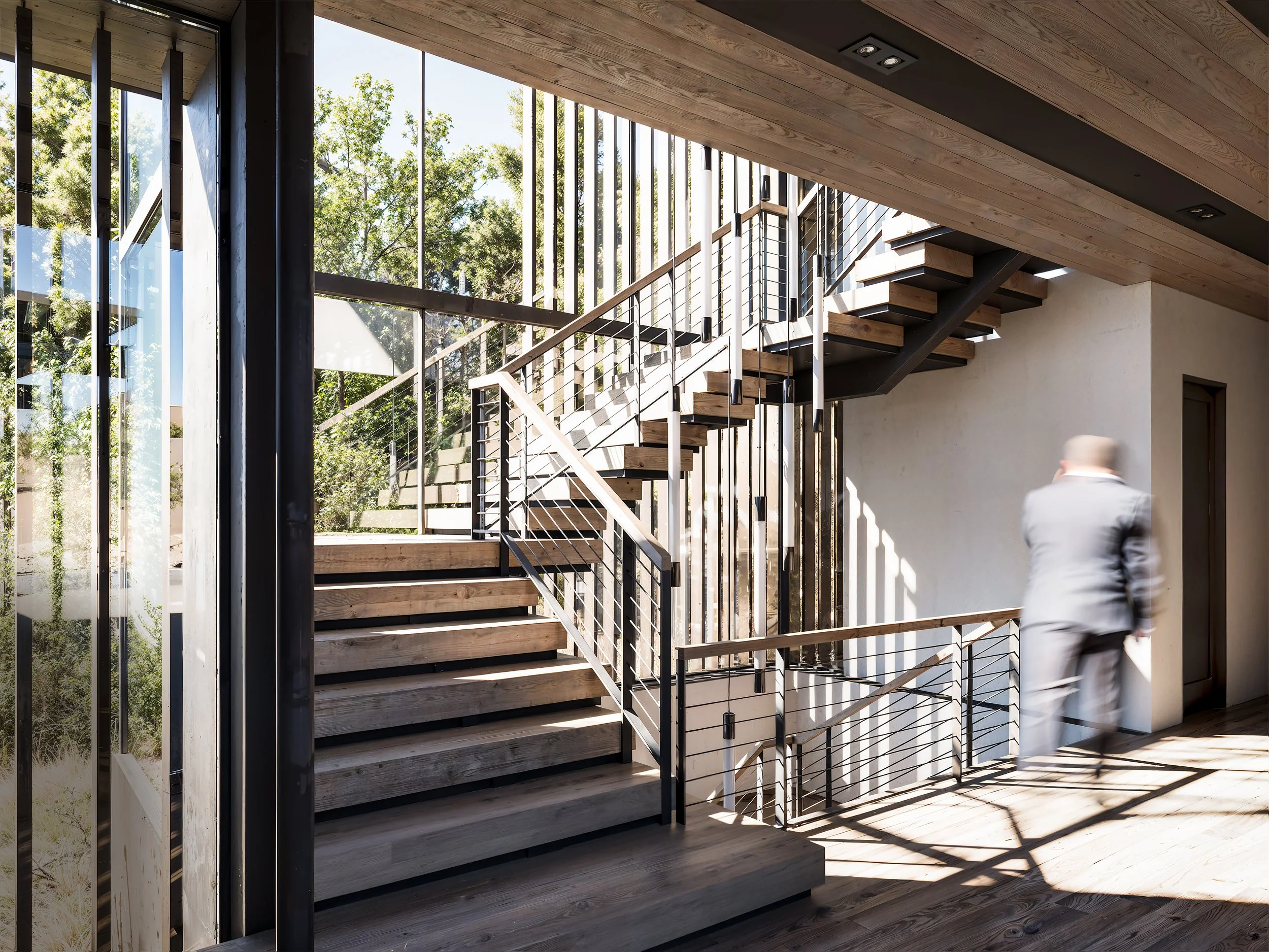 Modern interior with wooden stairs, large windows, and a man in a suit walking.