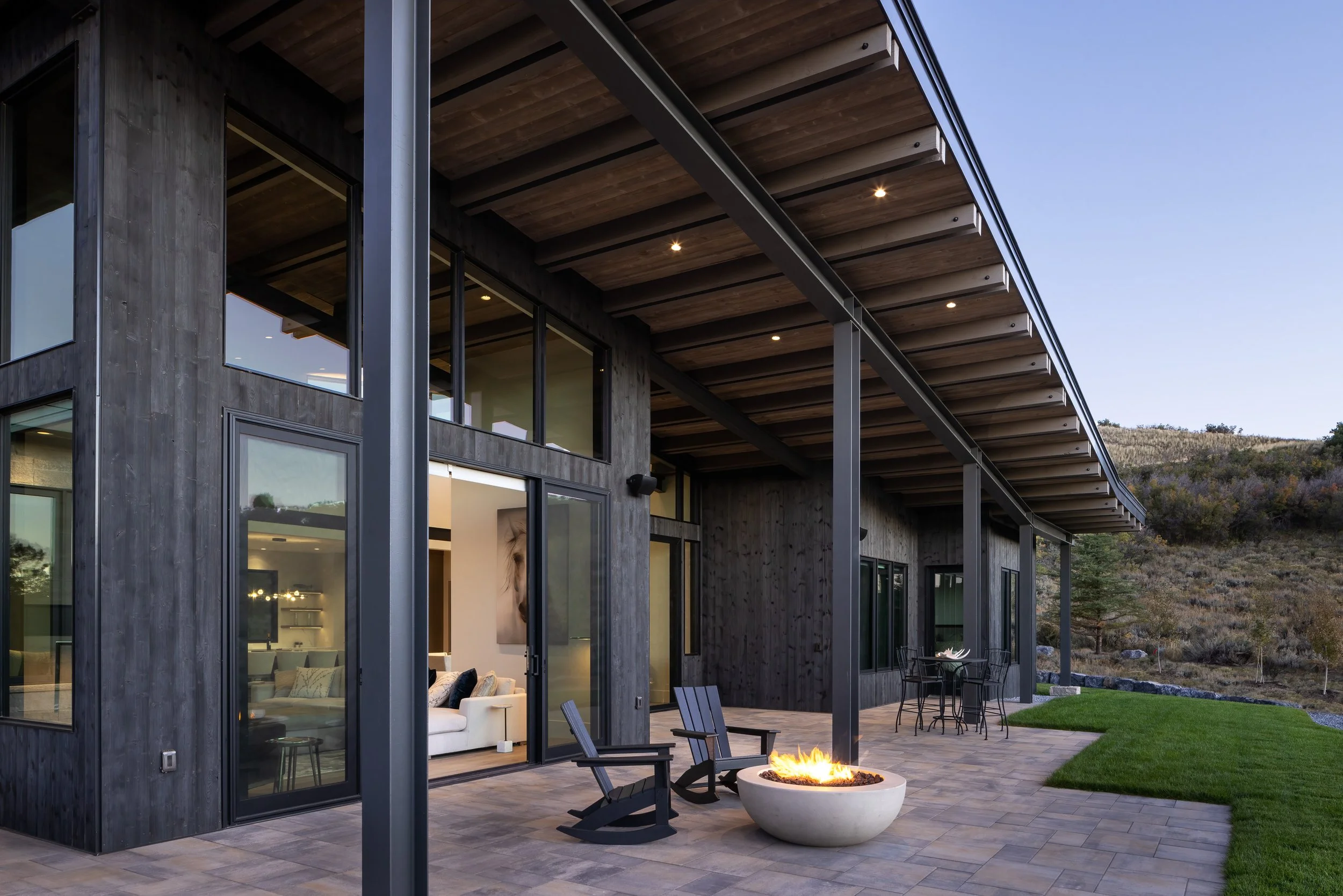 Modern house exterior with large glass windows and a patio featuring a fire pit, Adirondack chairs, and a dining table, set against a hilly landscape during dusk.