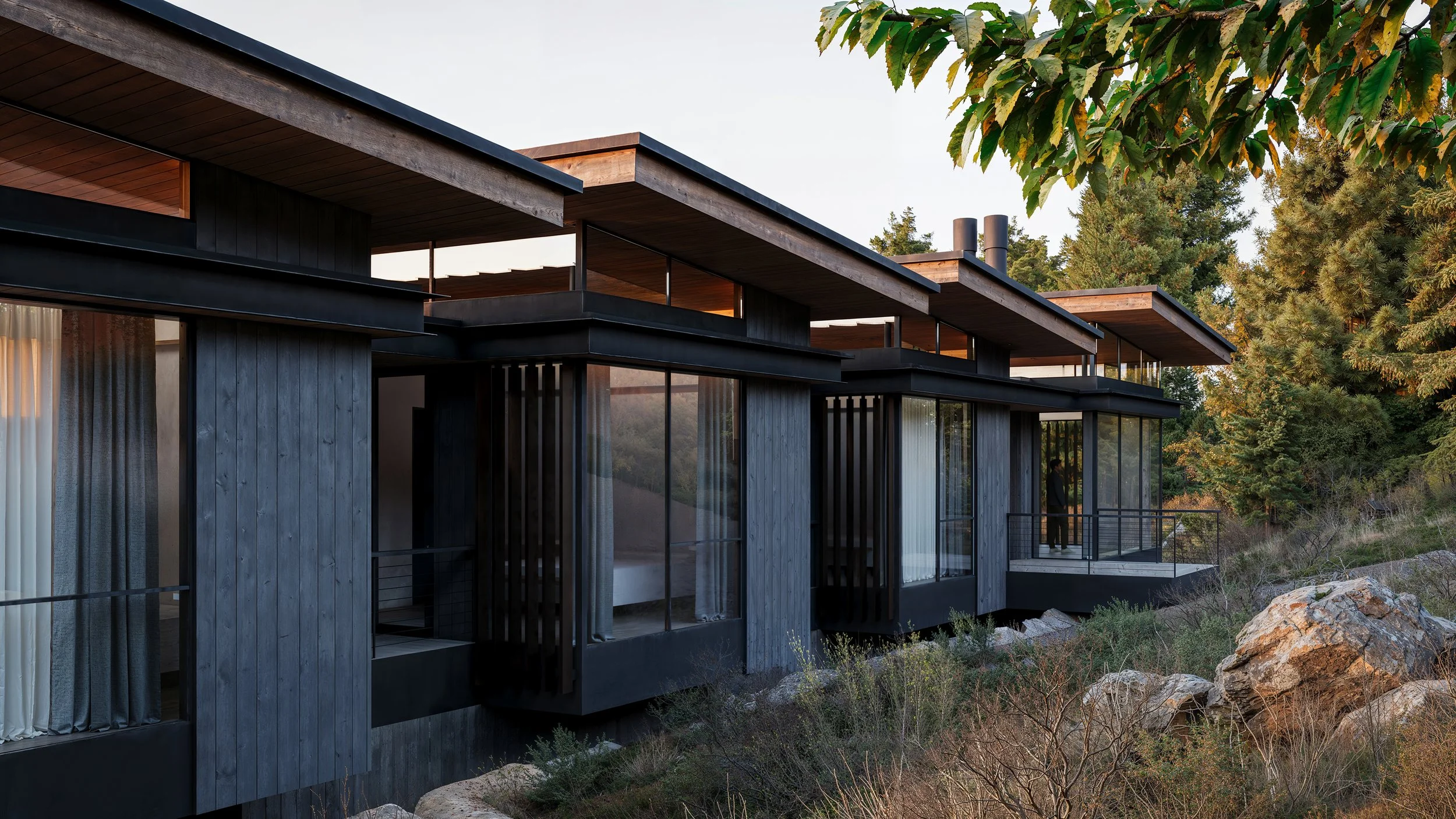 Modern house with large glass windows and wooden exterior, situated on a rocky hillside with surrounding trees, during sunset or late afternoon.