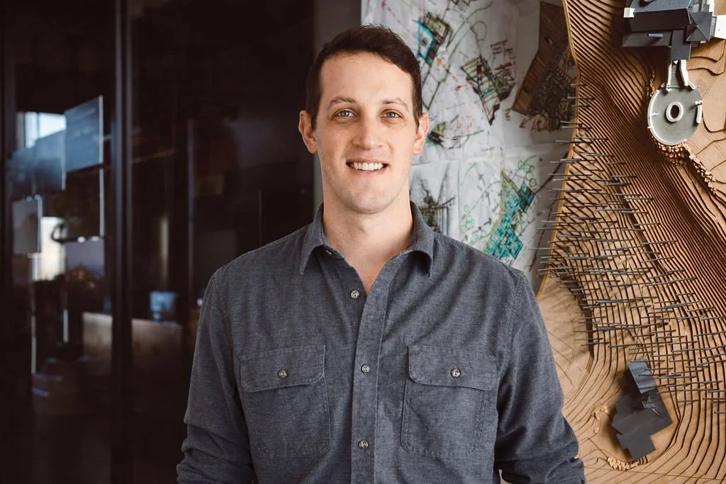A smiling man with short dark hair wearing a dark gray button-up shirt standing in front of a wall with abstract art and maps.