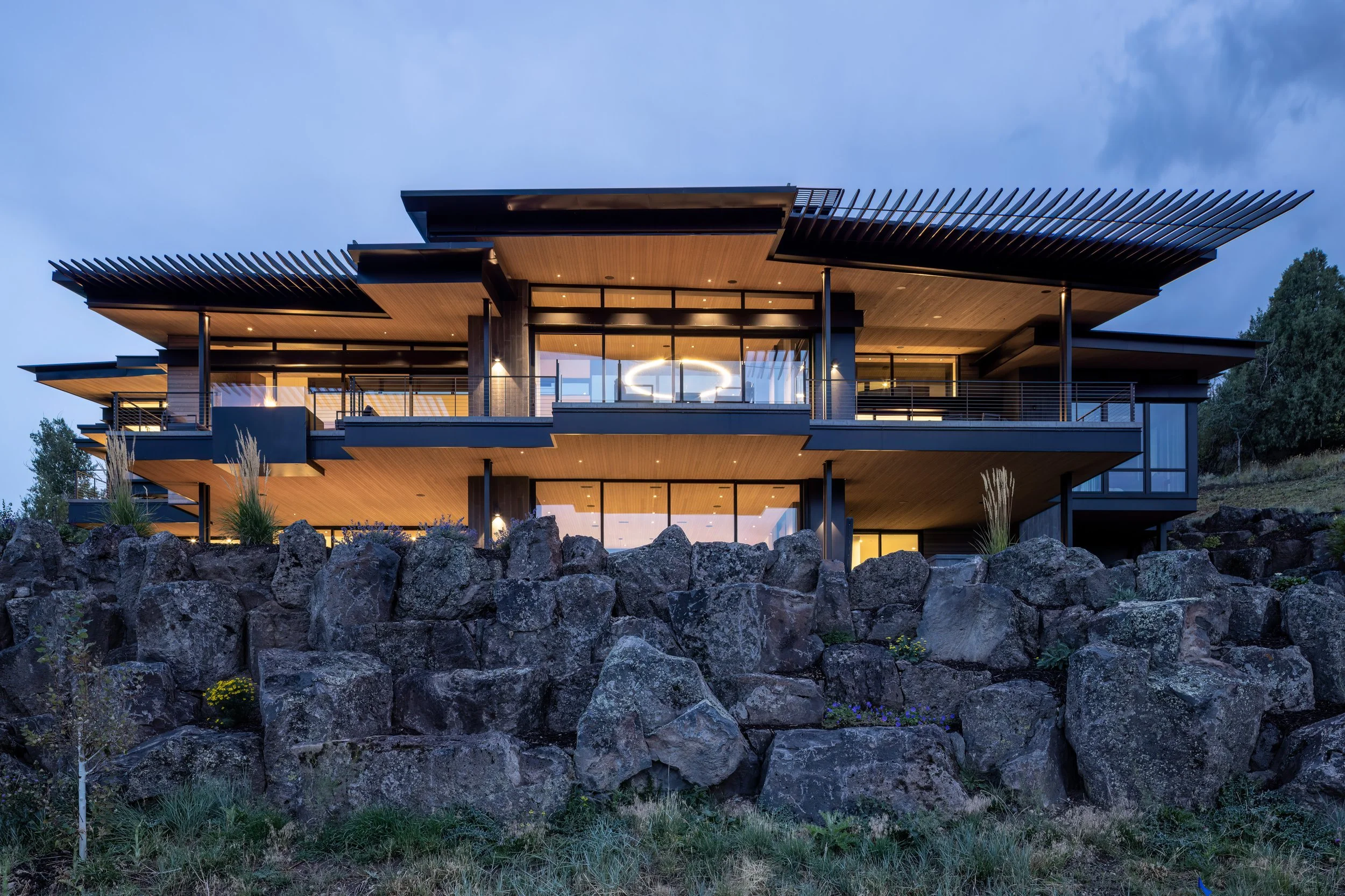 Modern multi-story house with large glass windows, wooden and black exterior design, located on rocky landscape, illuminated from within during dusk.
