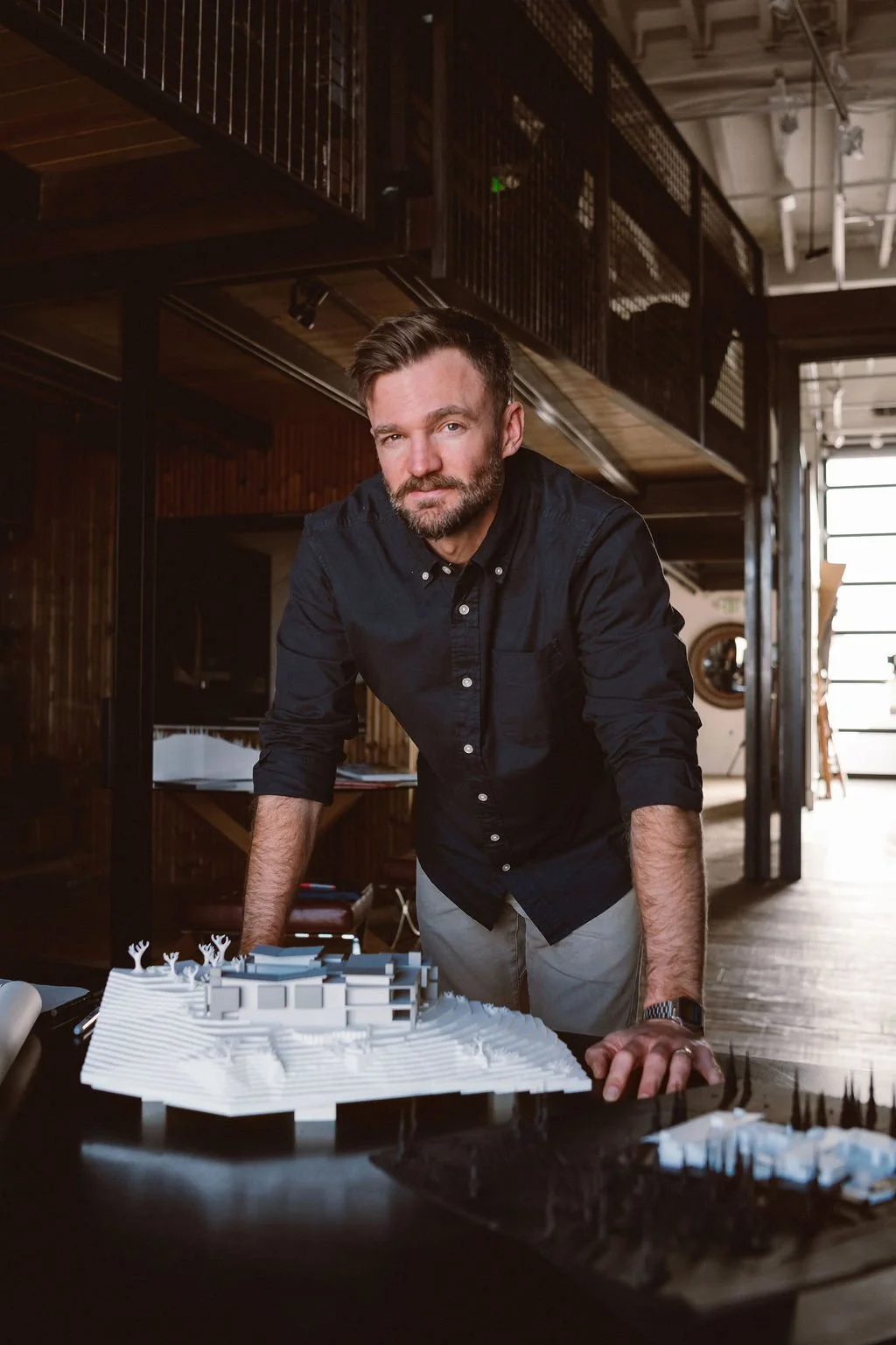 A man with a beard and short brown hair leaning on a table with architectural models in an industrial-style space with wooden walls and metal railings.