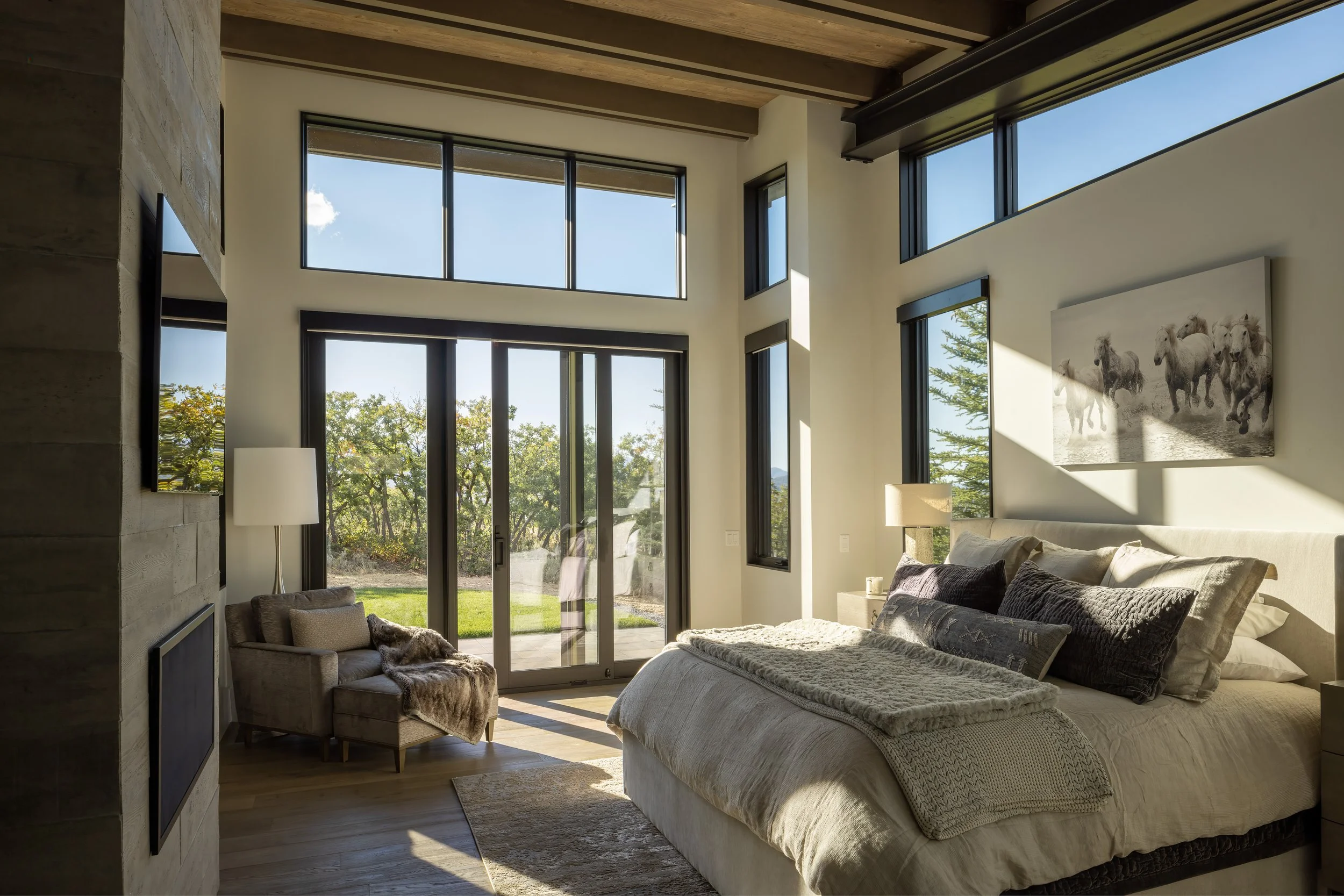 Bright bedroom with large windows, a bed with pillows, a small armchair with a throw blanket, and a landscape view of trees outside.