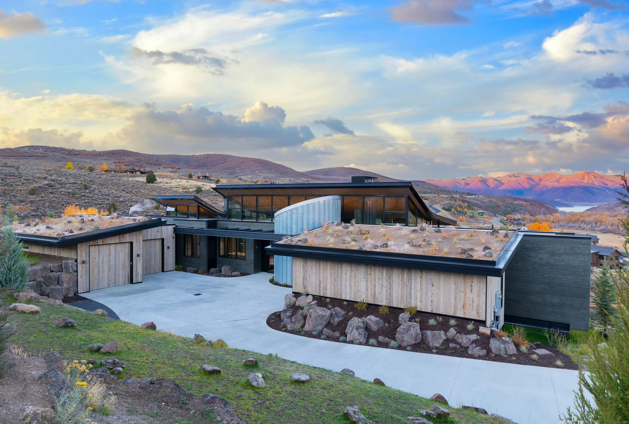 Modern house with flat roofs and extensive glass windows situated in a hilly landscape with mountains in the background, under a partly cloudy sky.
