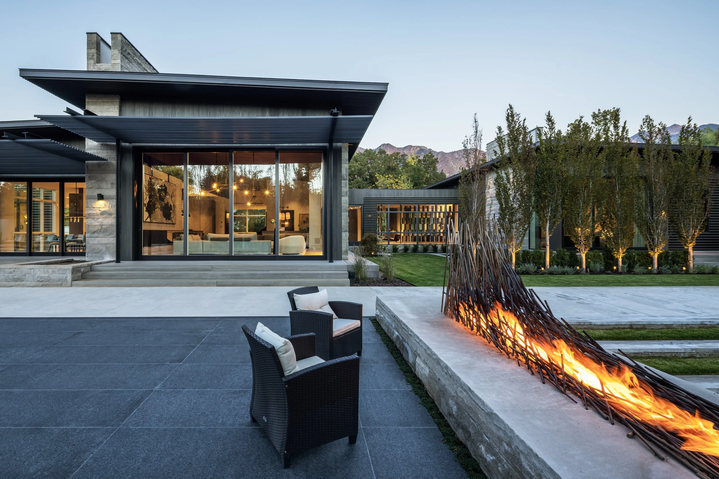 Modern house with large glass windows, outdoor fire feature, two black seating chairs with cushions, landscaped yard with trees, mountains in the background, and evening lighting.