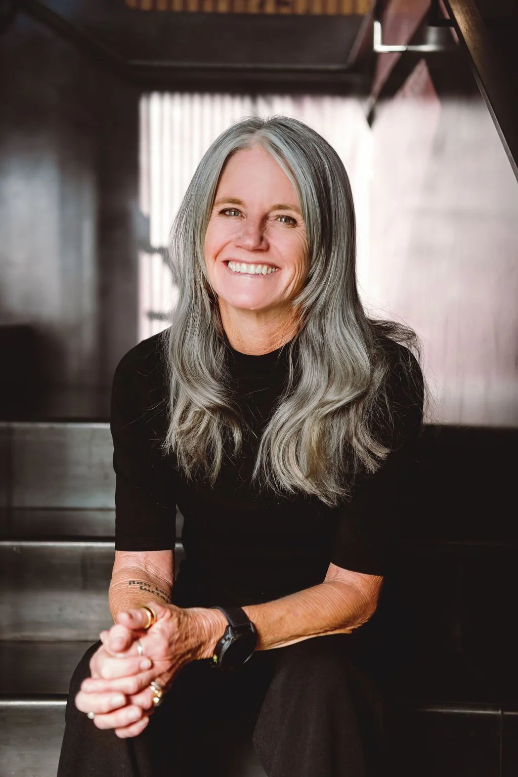 A smiling woman with long gray hair wearing a black top, sitting on stairs with blurred light background.