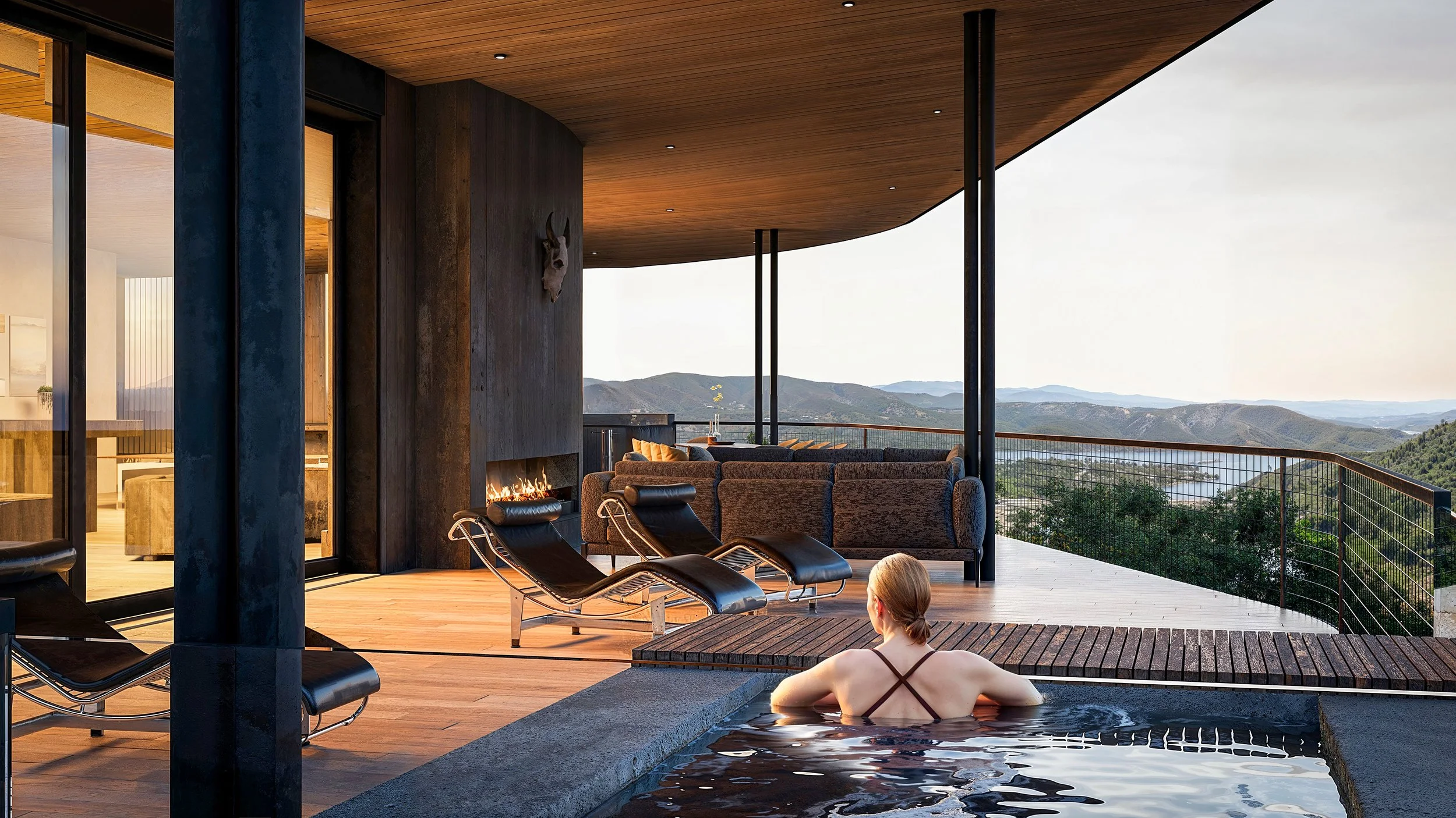 Woman relaxing in an outdoor hot tub with a view of rolling hills and mountains. Cozy outdoor seating and modern architecture with wood and glass elements.