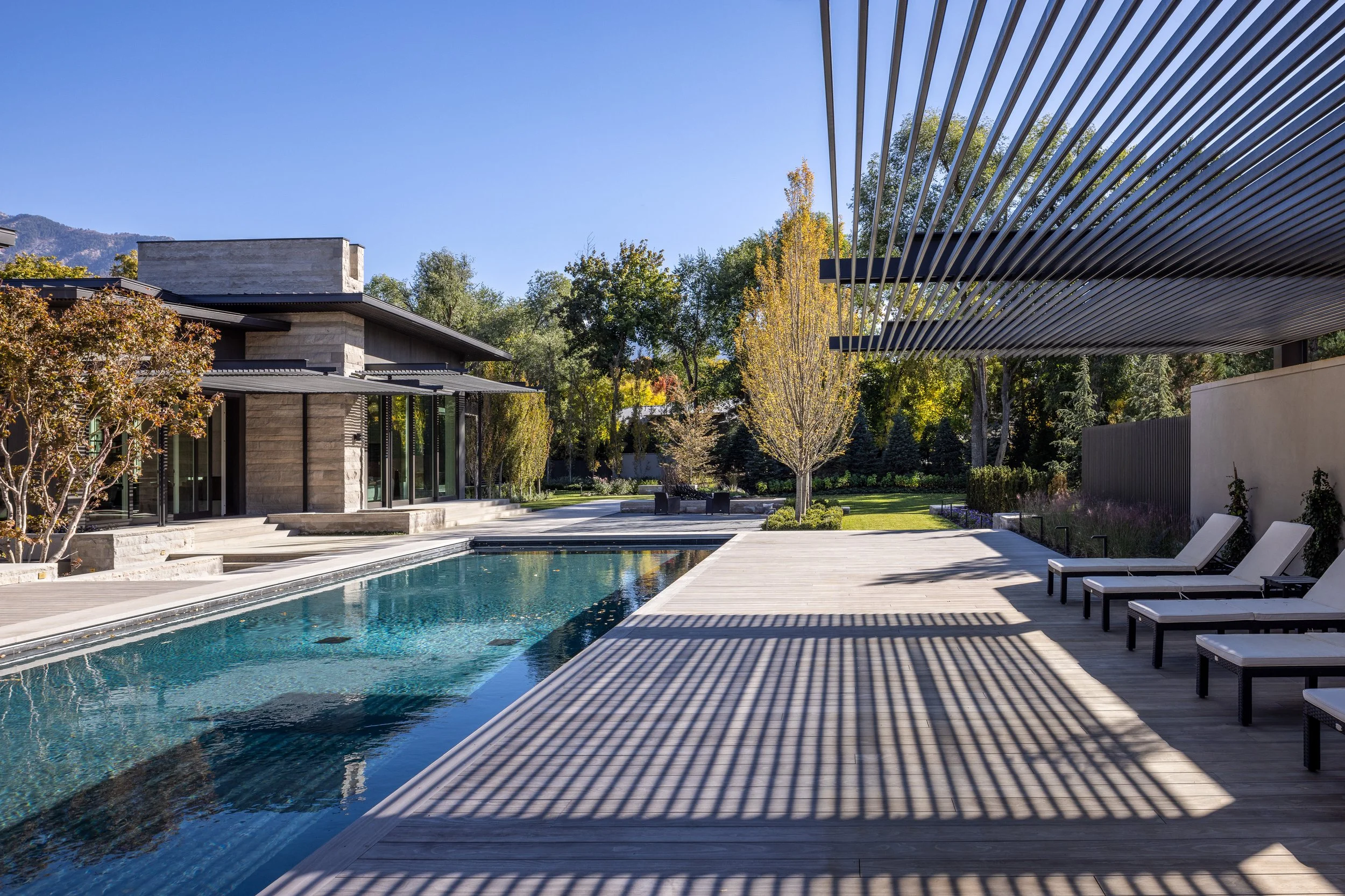 Modern backyard with swimming pool, lounge chairs, trees, and a house with large glass windows and metal awnings, under clear blue skies.
