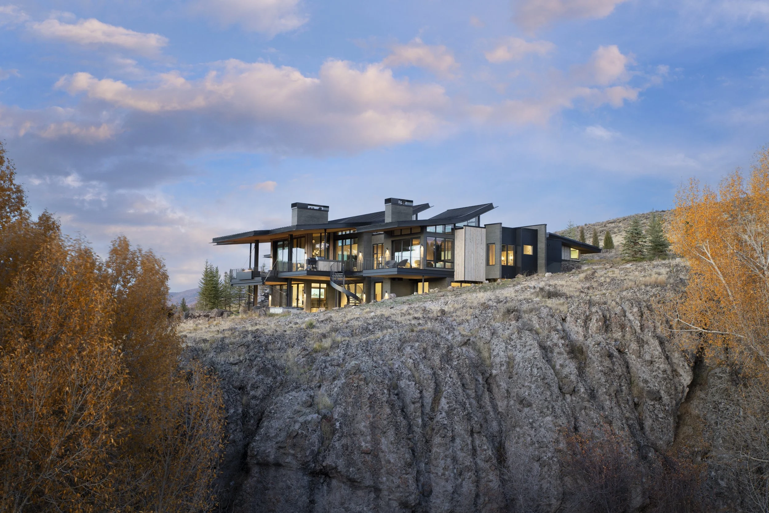 Modern house on a rocky hillside surrounded by autumn trees with a partly cloudy sky in the background.