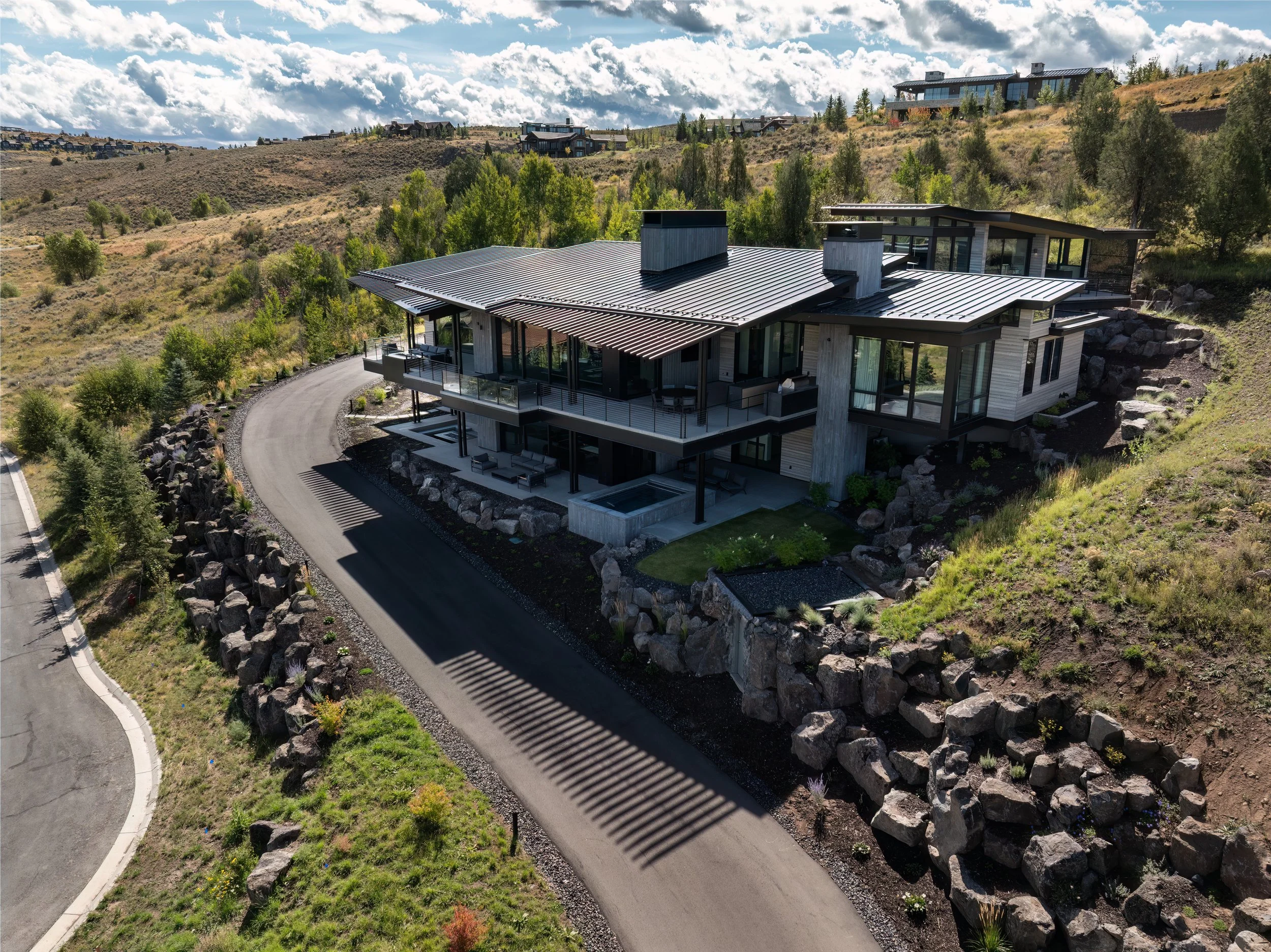 A modern house on a hillside with large windows, multiple balconies, and a sloped metal roof, surrounded by rocks, trees, and a winding driveway under a partly cloudy sky.