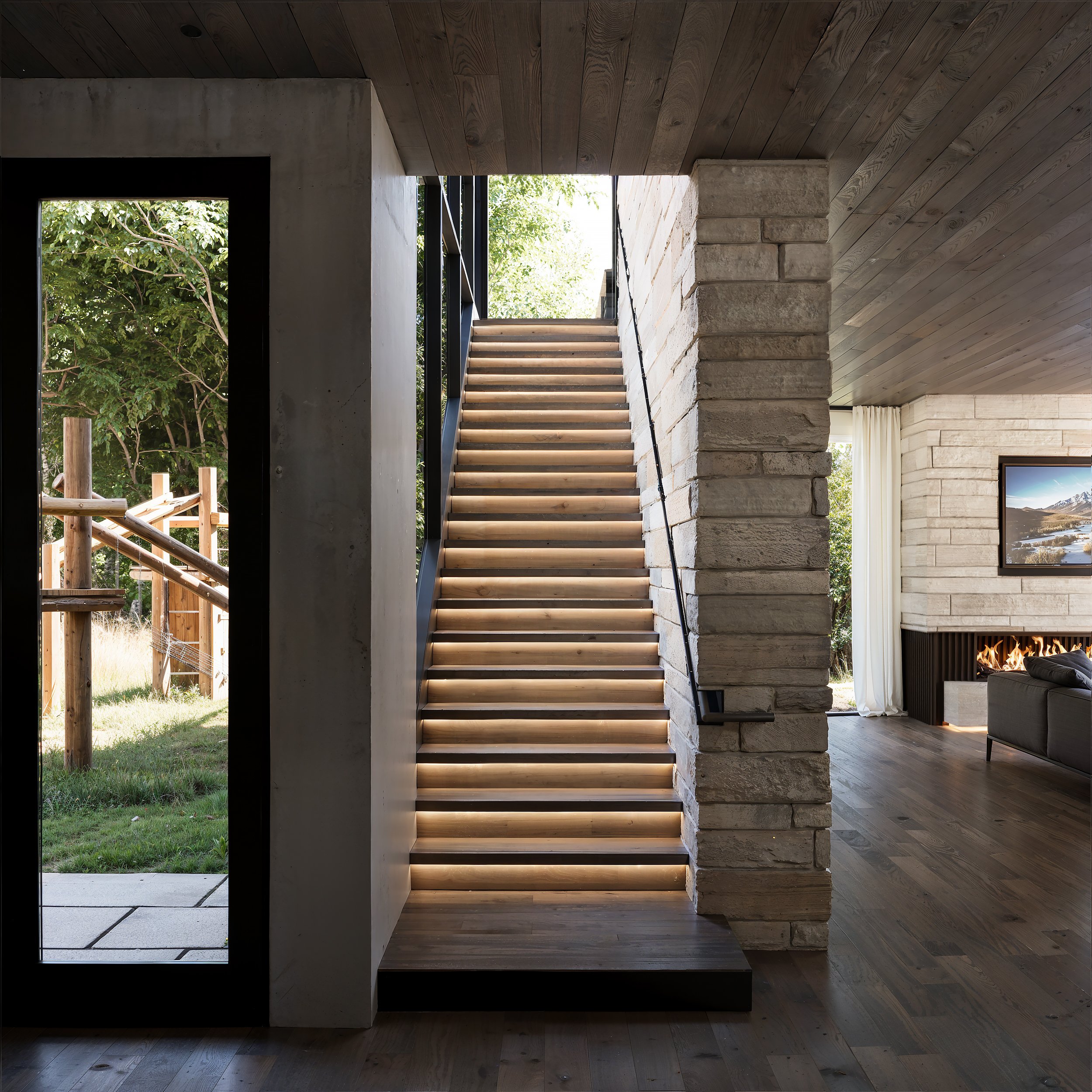 Interior view of a modern home with a staircase, stone and wood accents, and a living room with a fireplace and a wall-mounted TV.