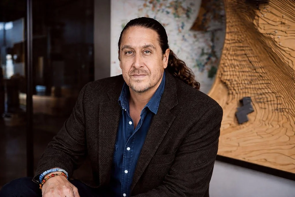 Portrait of a middle-aged man with long dark hair and light beard, wearing a dark blazer and blue shirt, sitting indoors with abstract art behind him.