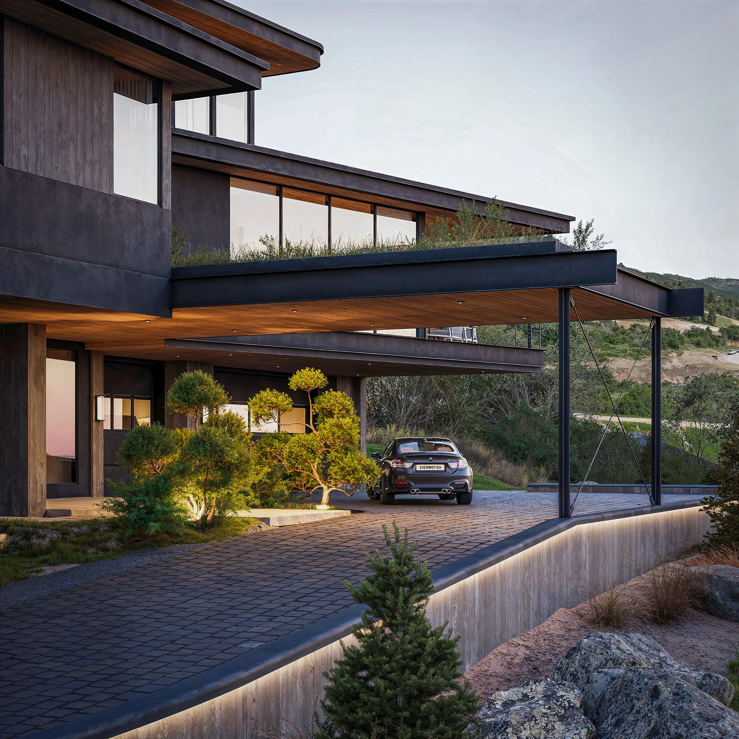 Modern multi-story house with large windows, wooden accents, and outdoor patio, surrounded by landscaped greenery and rocks, with a black sports car parked in driveway during the daytime.