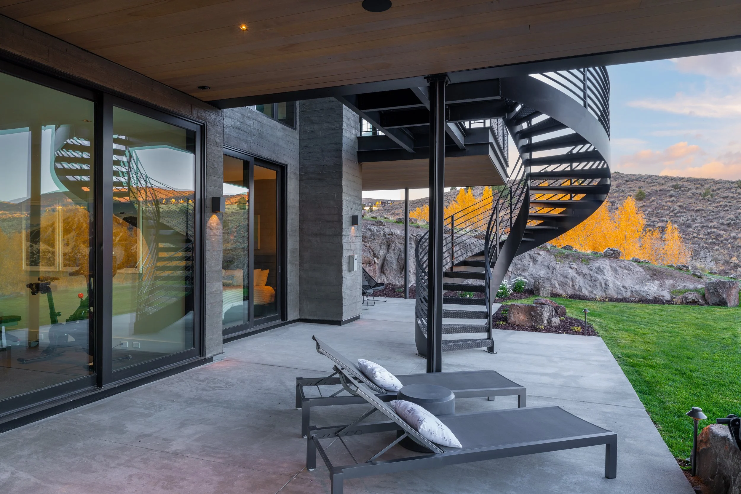 Modern outdoor patio with two lounge chairs, a coiled metal staircase, sliding glass doors, and landscaped yard with rocks and trees in the background during sunset.