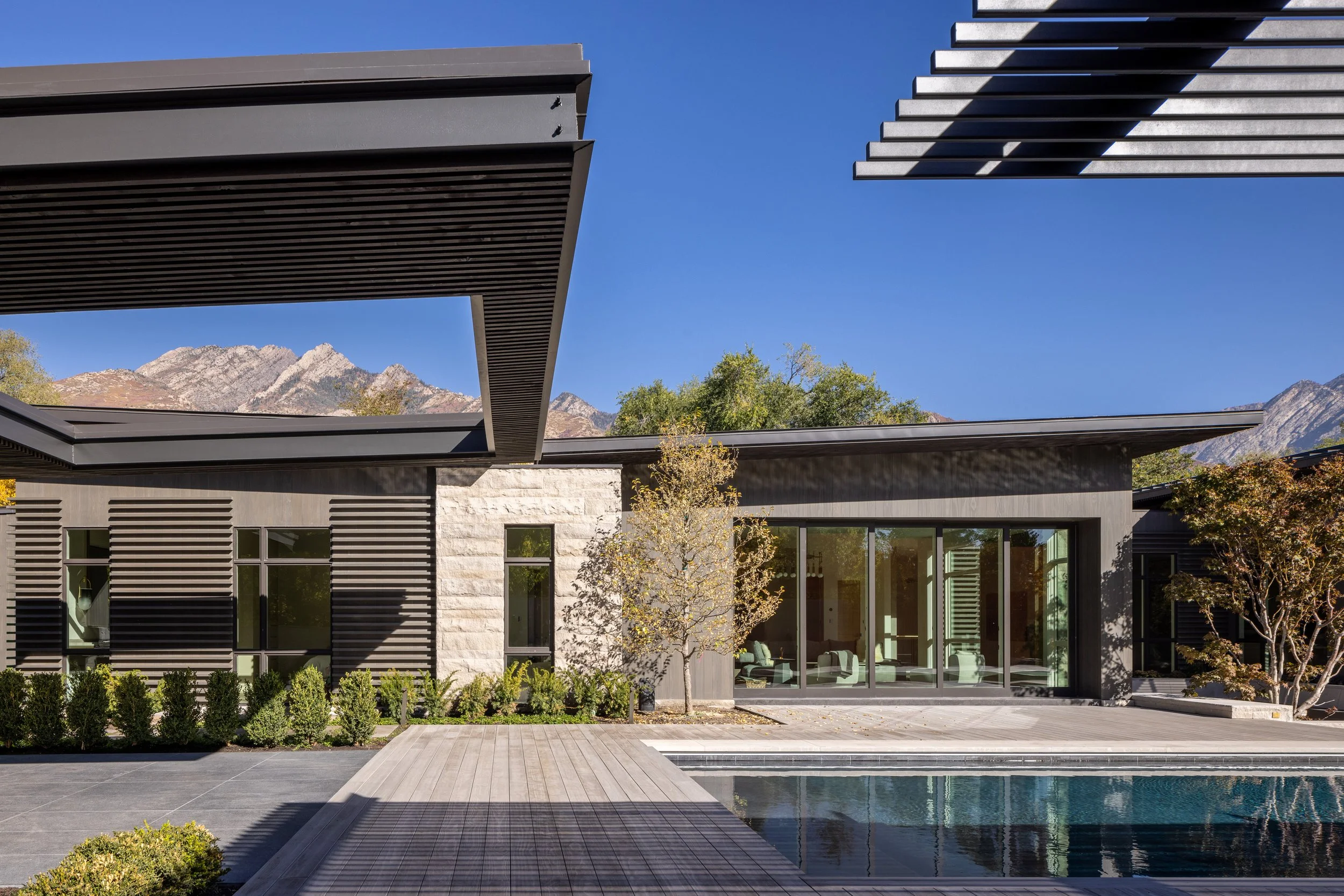 Modern house with large glass windows, surrounded by a backyard with a swimming pool, trees, and mountains in the background under a clear blue sky.