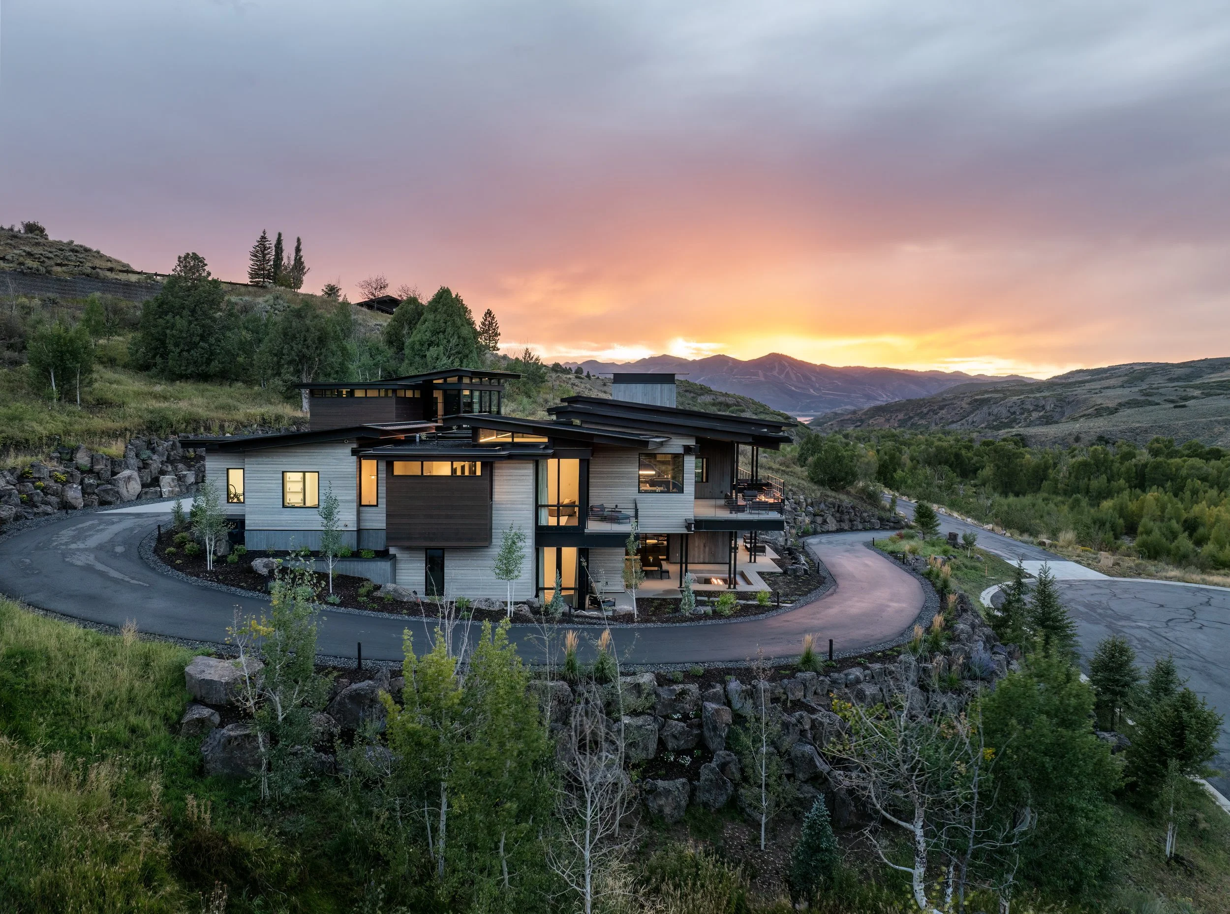 Modern house with multiple levels, large glass windows, and dark roofing, built into a hillside, during sunset with a mountain range in the background.