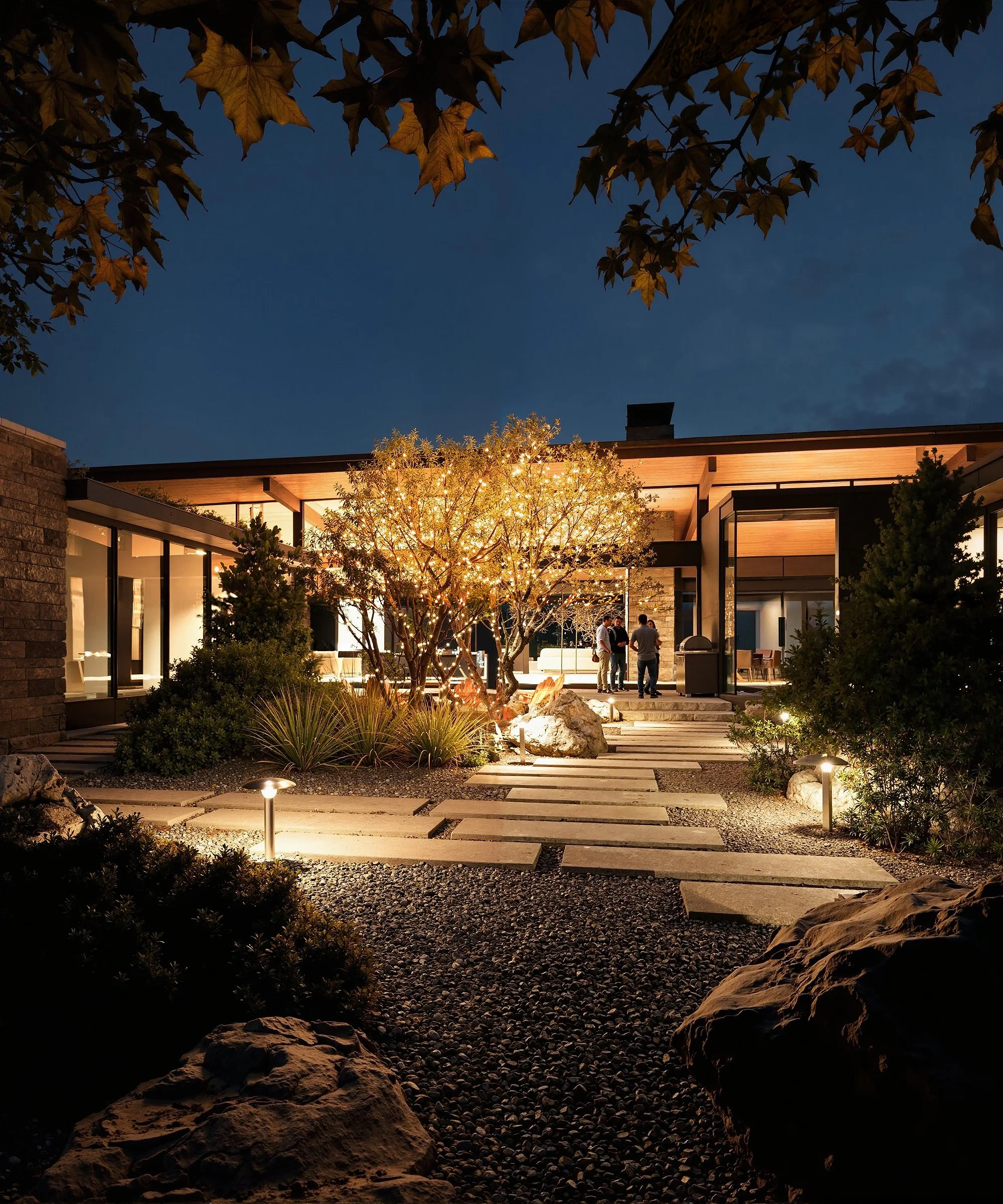 Modern house with large glass windows illuminated at night, surrounded by a landscaped garden with pathway lights, trees, bushes, and rocks.