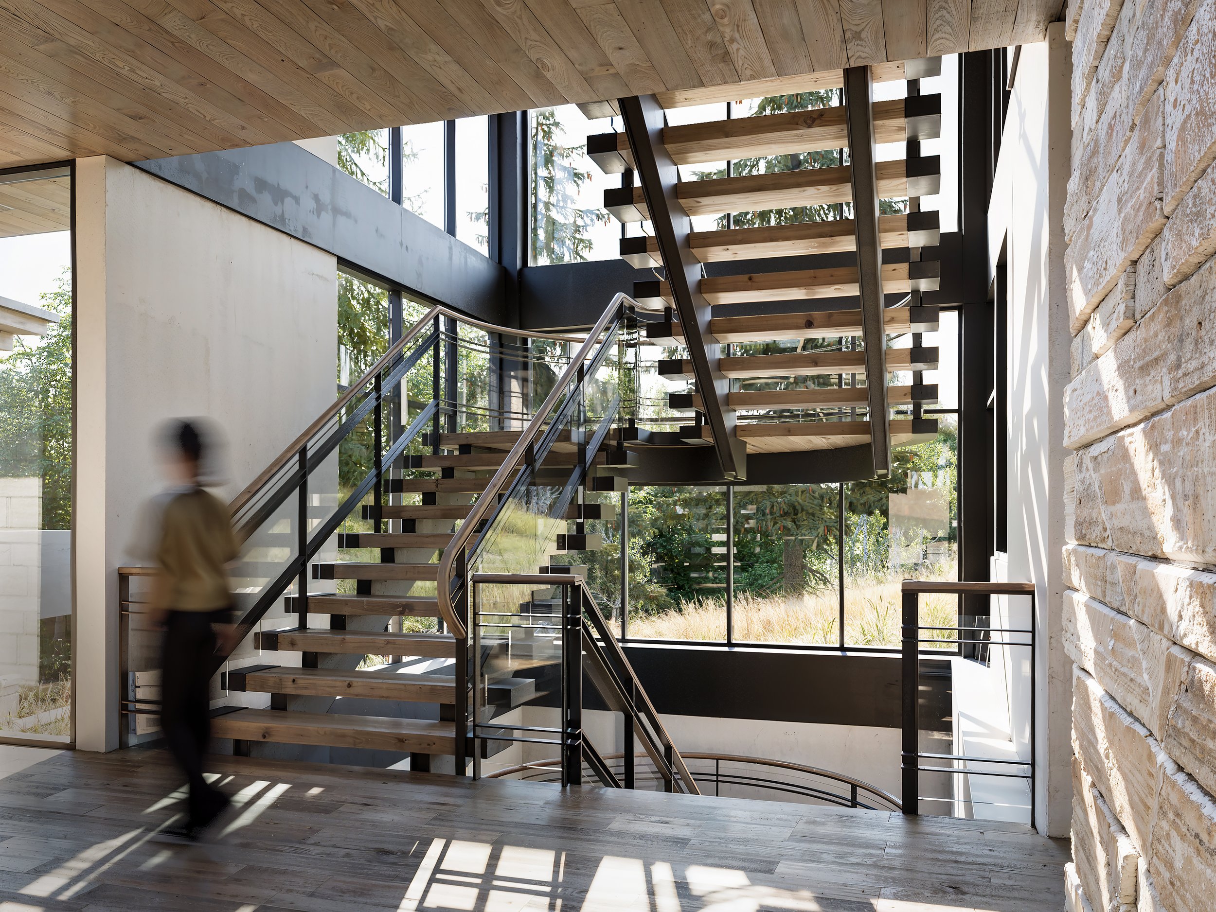 Interior view of a modern building staircase with wooden steps, black railings, and large windows showing greenery outside.