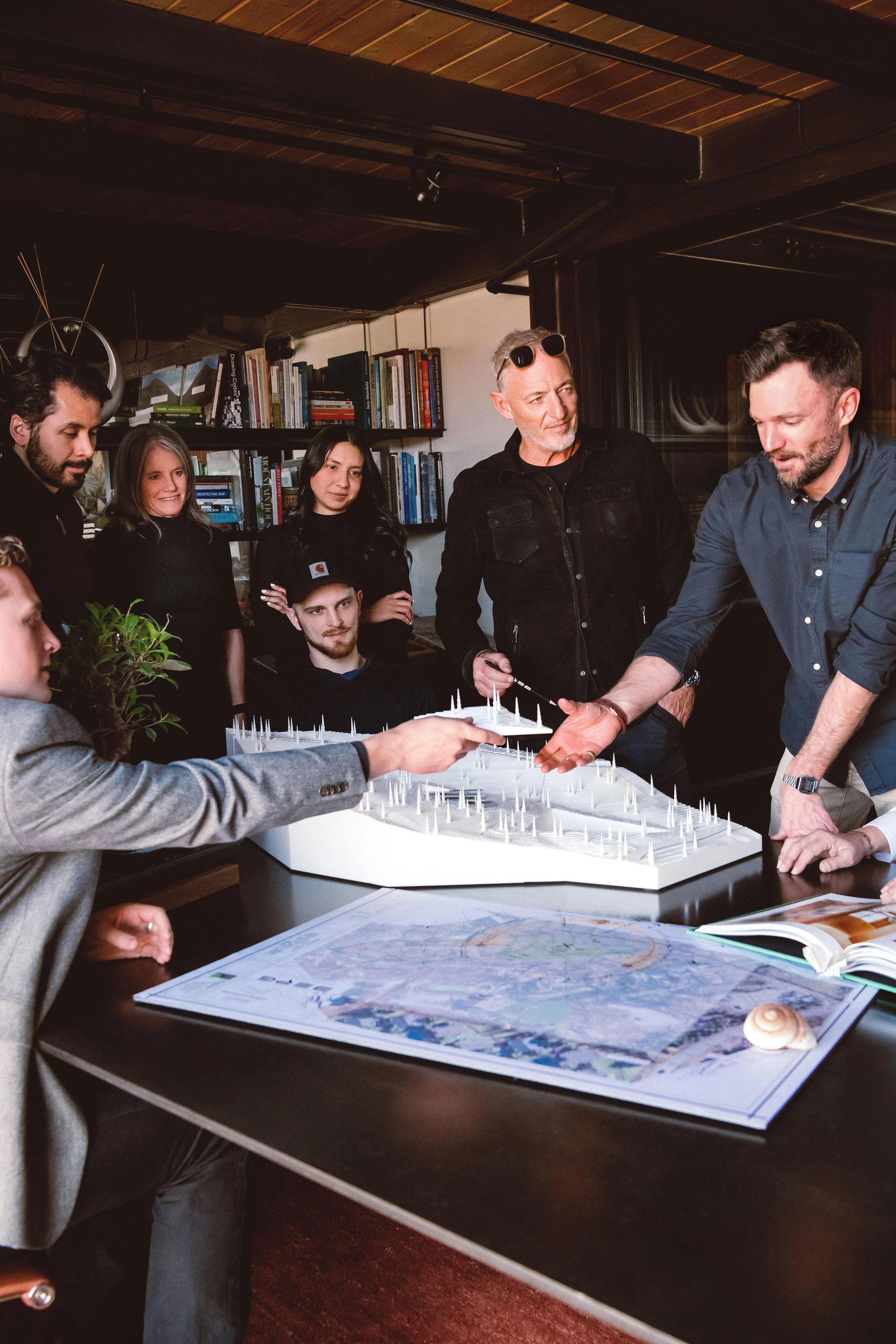 Group of people gathered around a table with a scale model, maps, and documents, in a room with bookshelves and wooden ceiling.