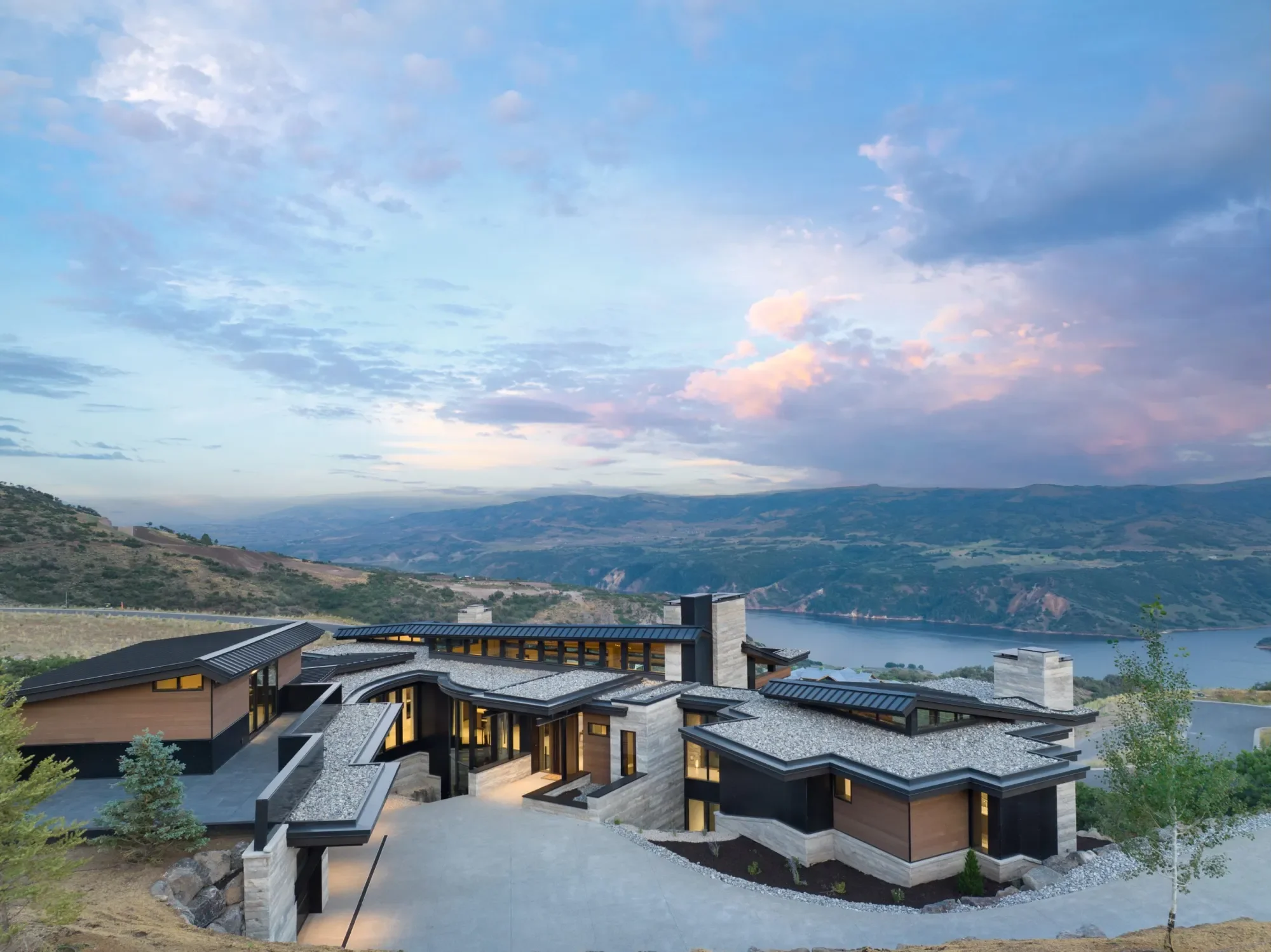 Modern house with multiple flat and sloped roofs near a lake, surrounded by hills and mountains under a partly cloudy sky at dusk.