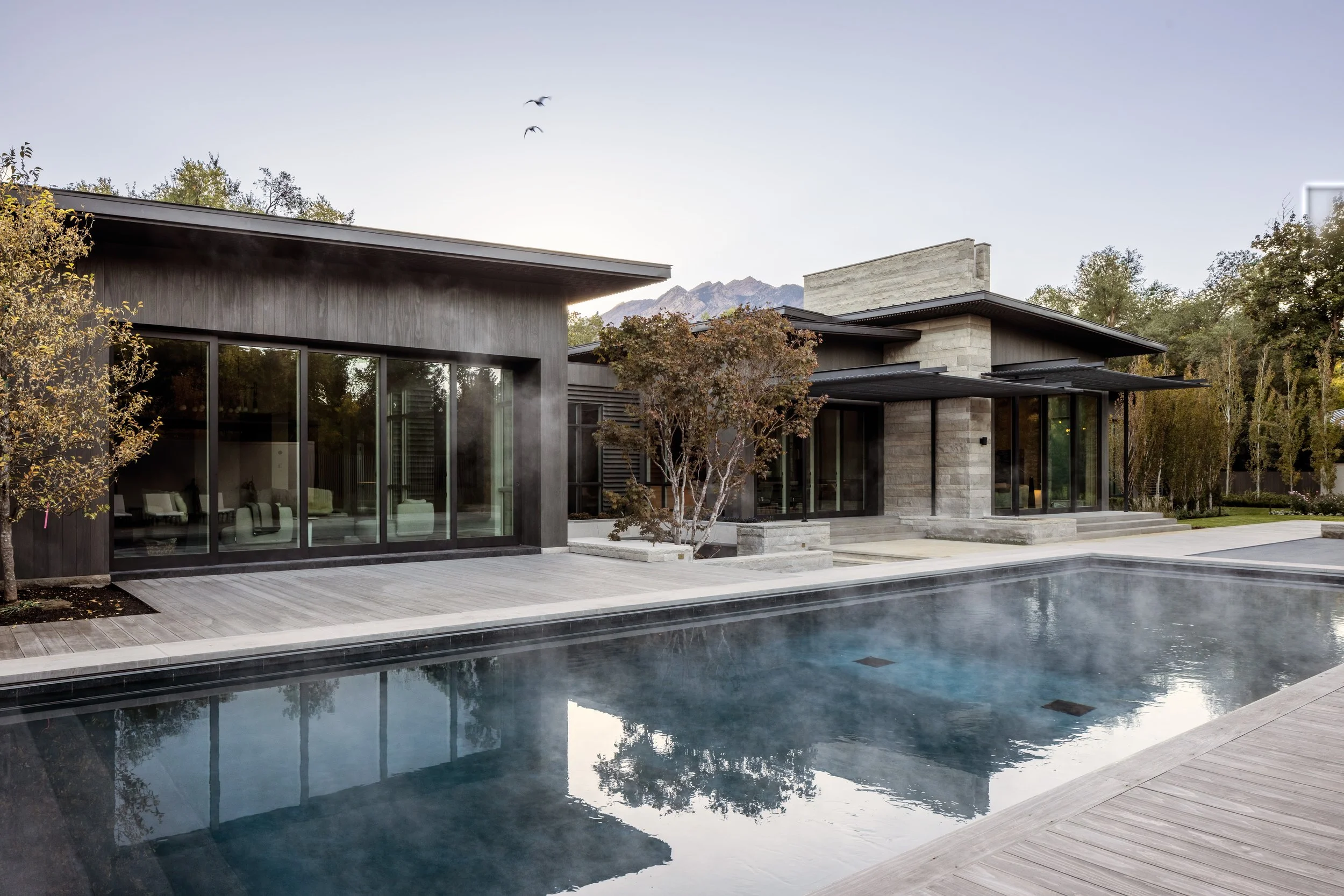 Modern house with large glass windows, wooden deck, a swimming pool, and trees in the yard, mountains in the background.