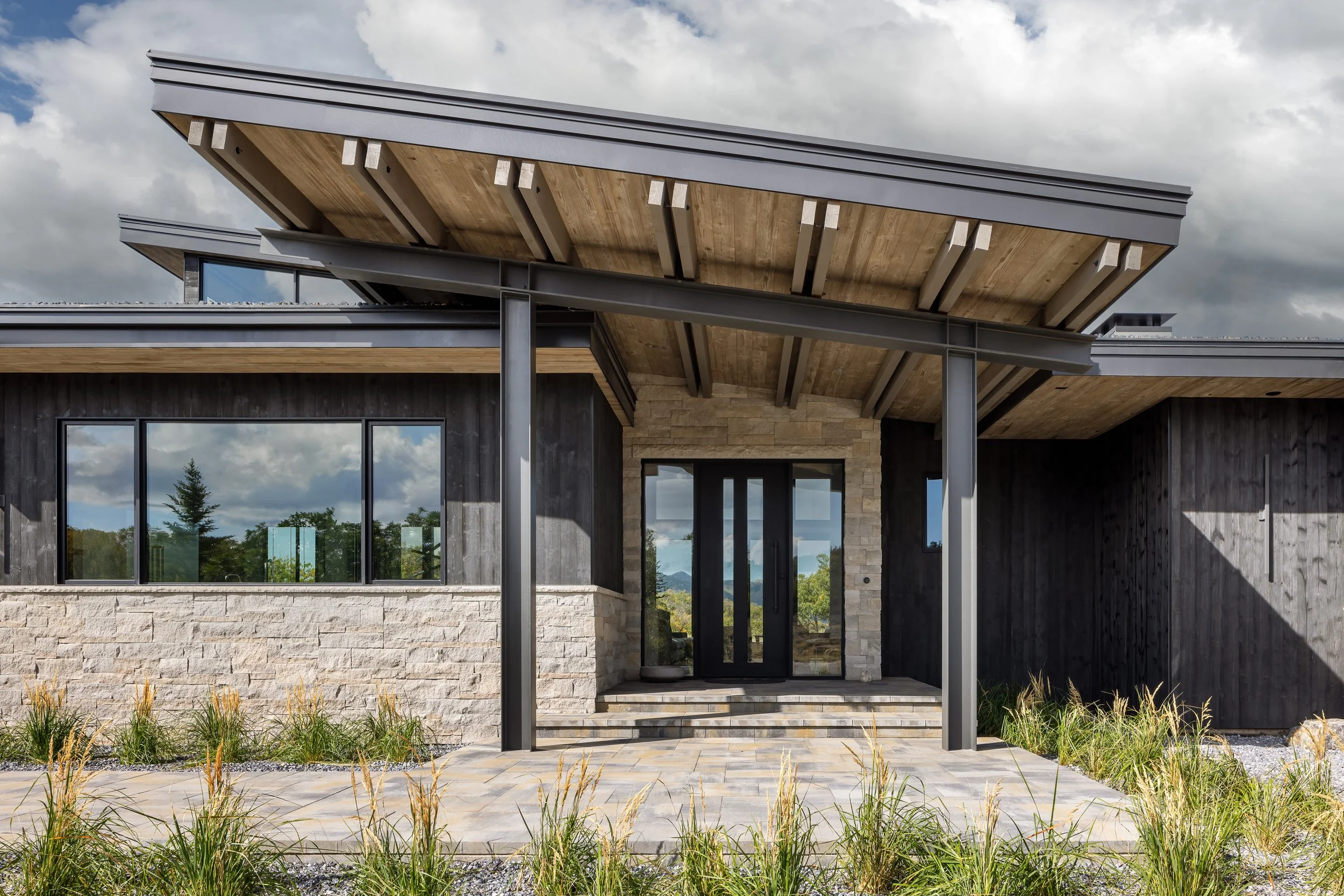 Modern house exterior with large glass windows, stone and wood siding, and a covered porch with steps, set against a partly cloudy sky.