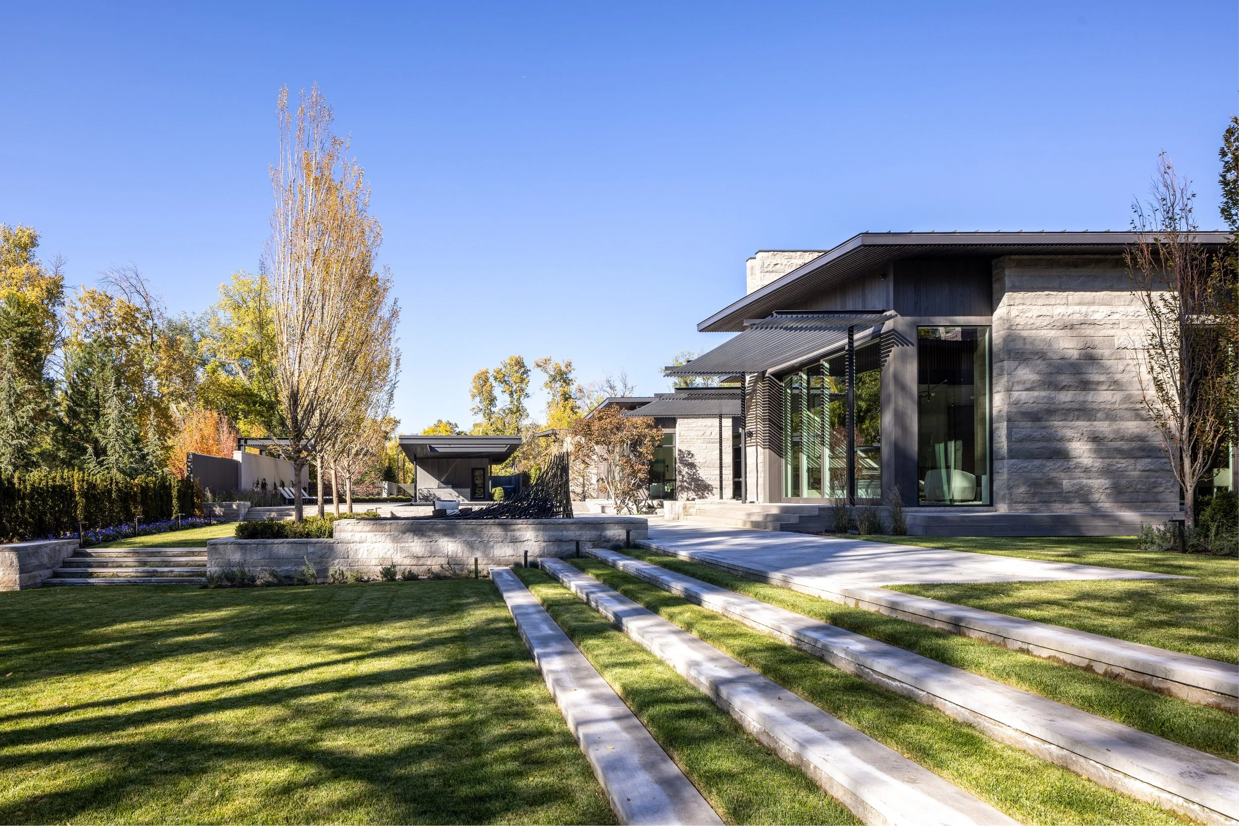 Modern house with large glass windows and surrounding landscaped yard with trees and grass under a clear blue sky.
