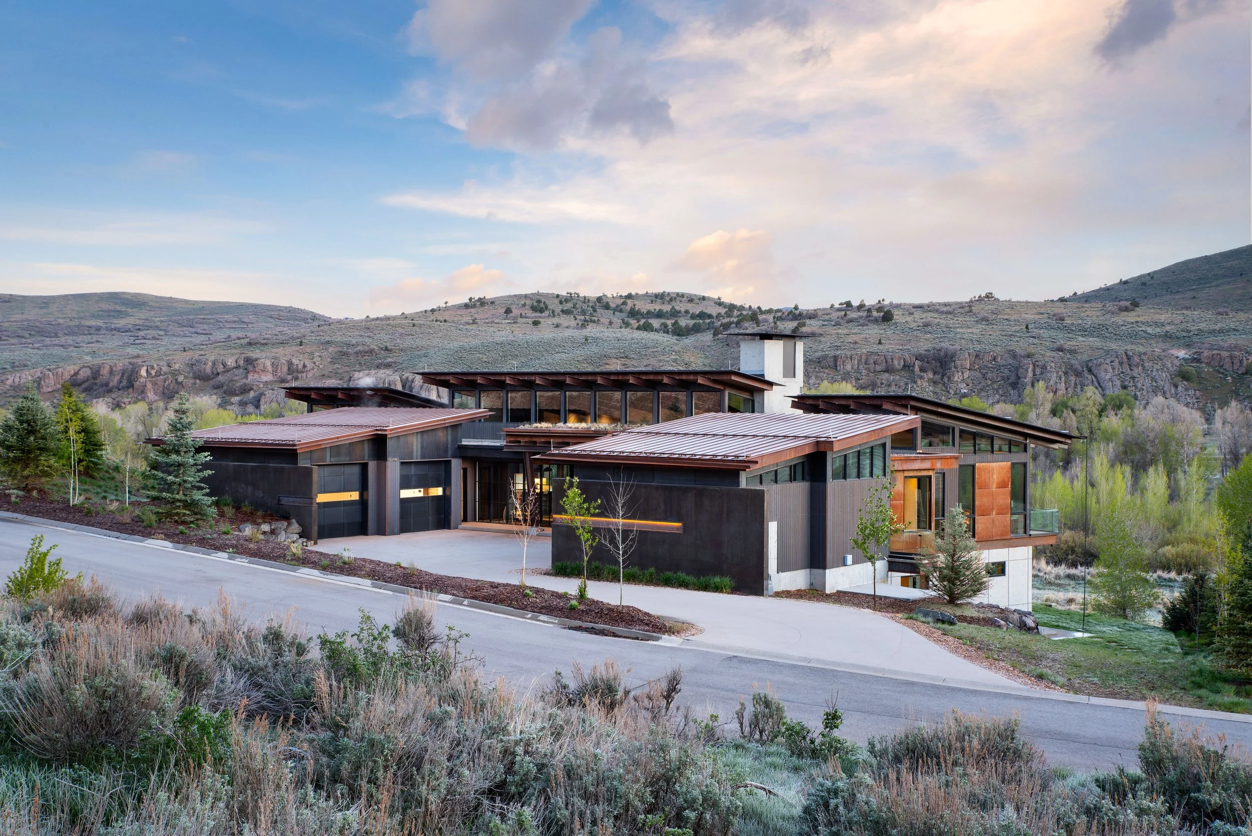 Modern house with black walls and large glass windows on a sloped landscape, surrounded by trees and mountains, under a partly cloudy sky.