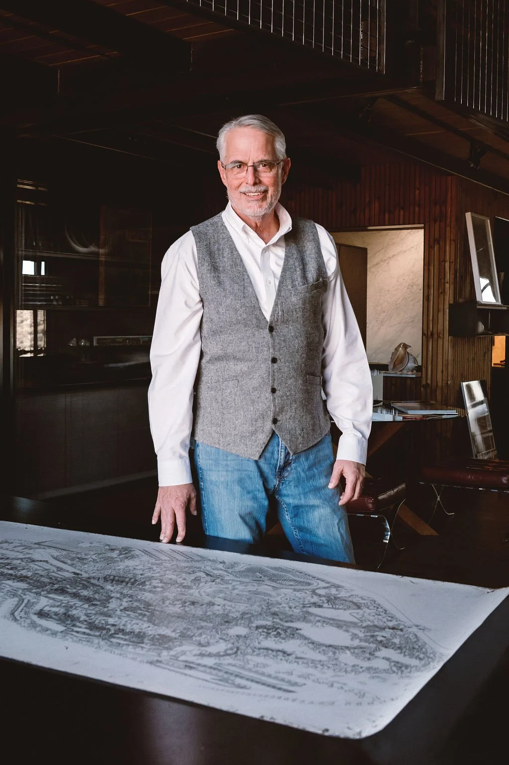 A senior man with gray hair and glasses standing in a room with wooden walls, smiling at the camera, dressed in a white shirt, gray vest, and jeans, with a large rolled-out architectural blueprint on a table in front of him.