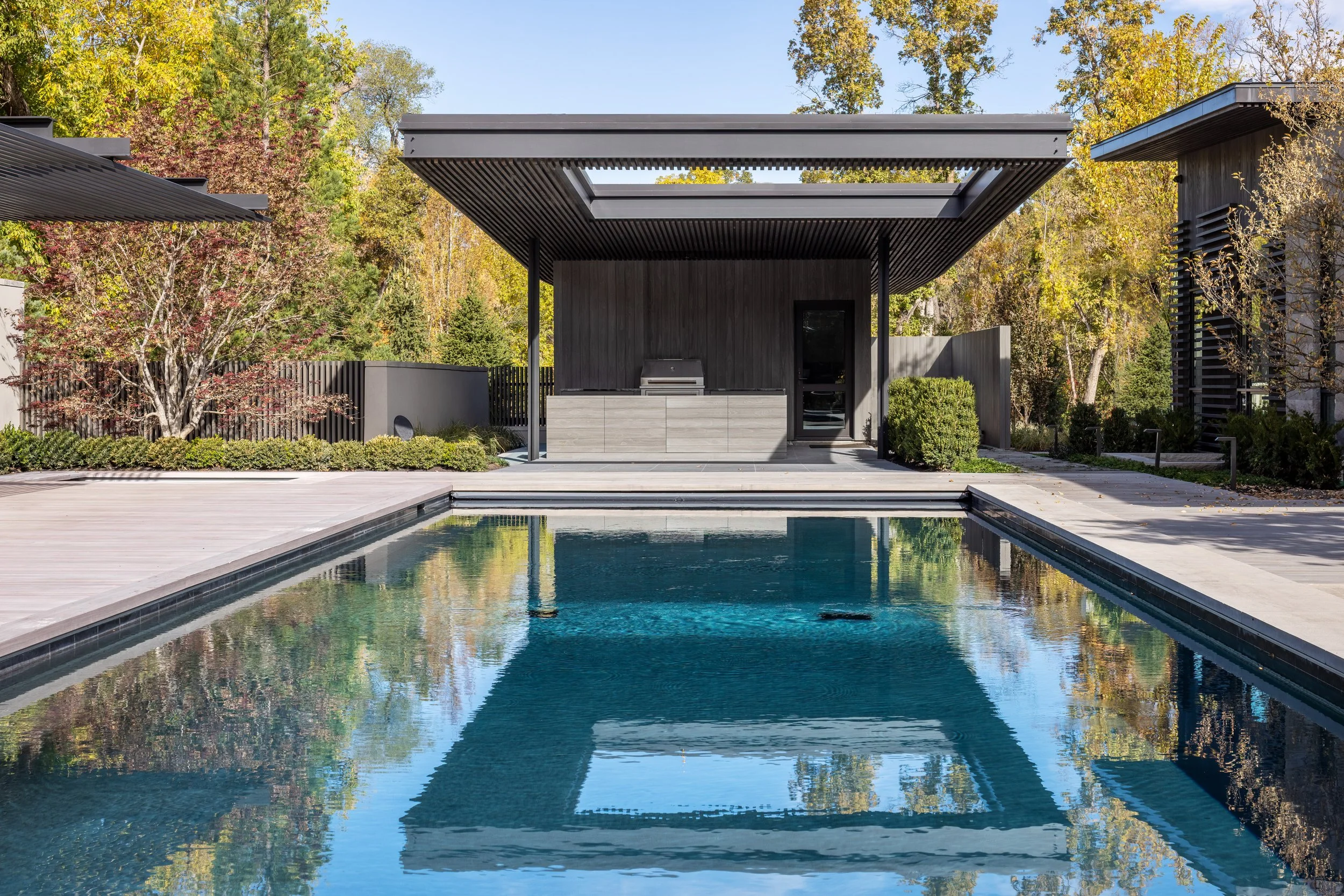 Modern backyard with a swimming pool, a covered outdoor kitchen area with grill, and surrounding greenery.